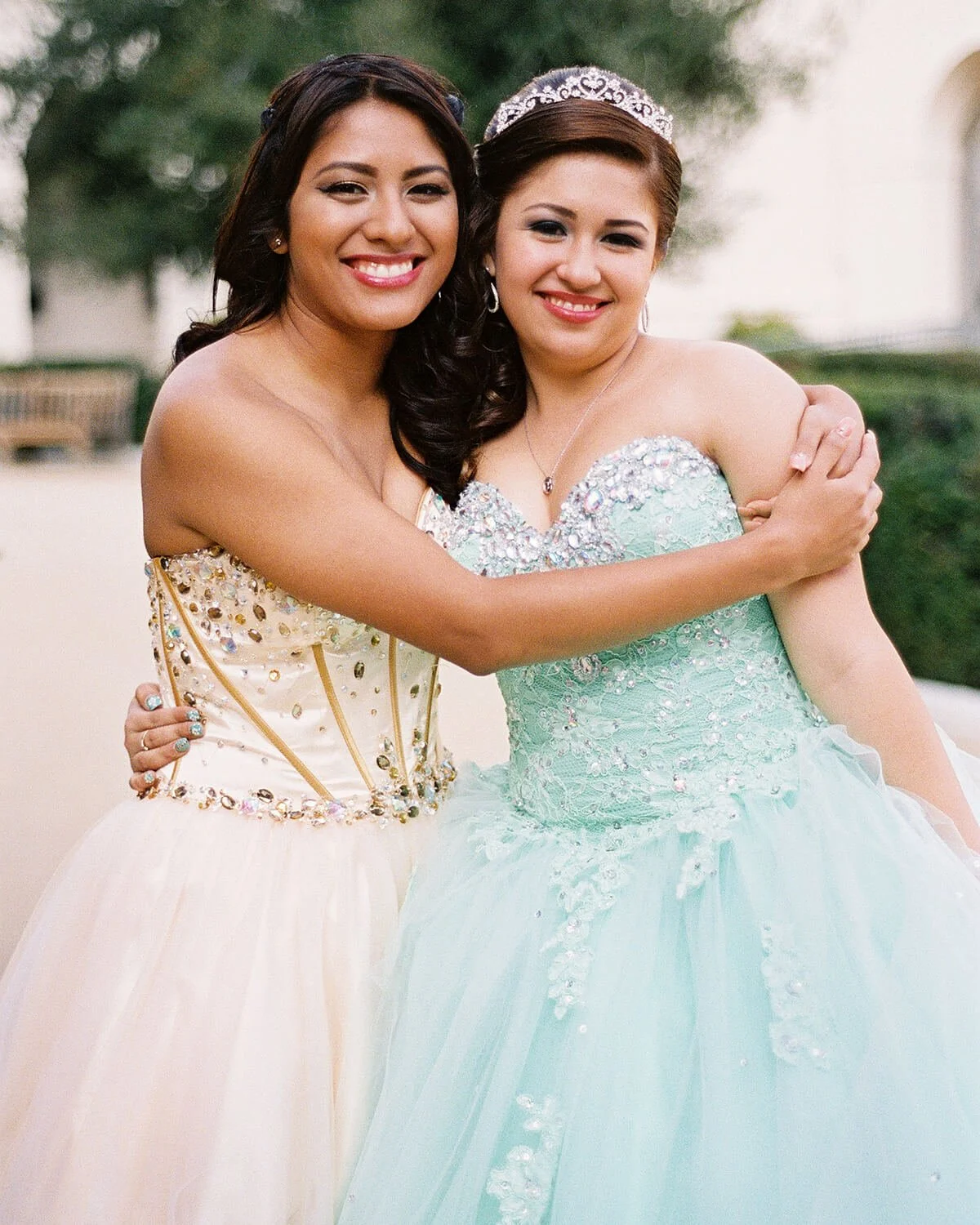 Two women in formal Quinceañera dresses hugging and smiling outdoors.