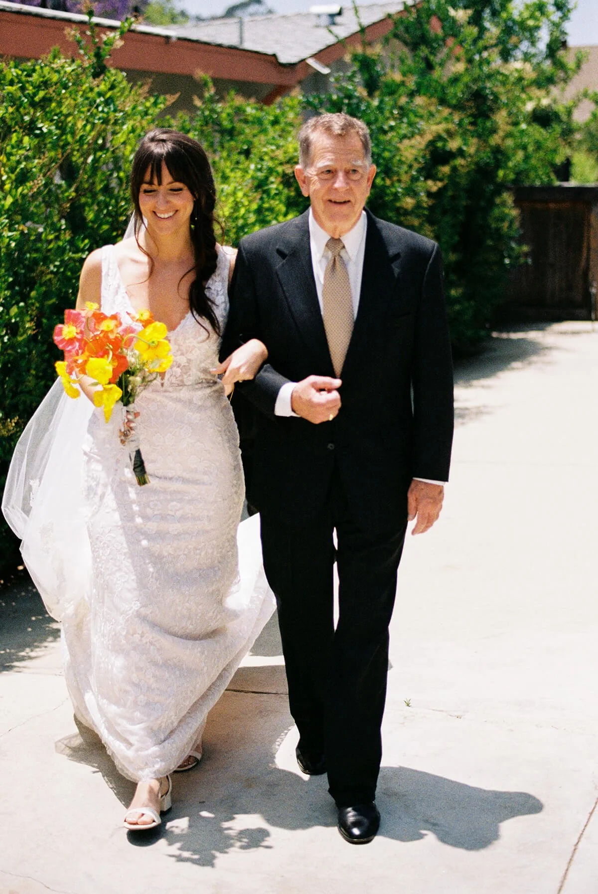 A bride in a white wedding dress holding a colorful bouquet walking arm-in-arm with an older man in a black suit on a sunny sidewalk.