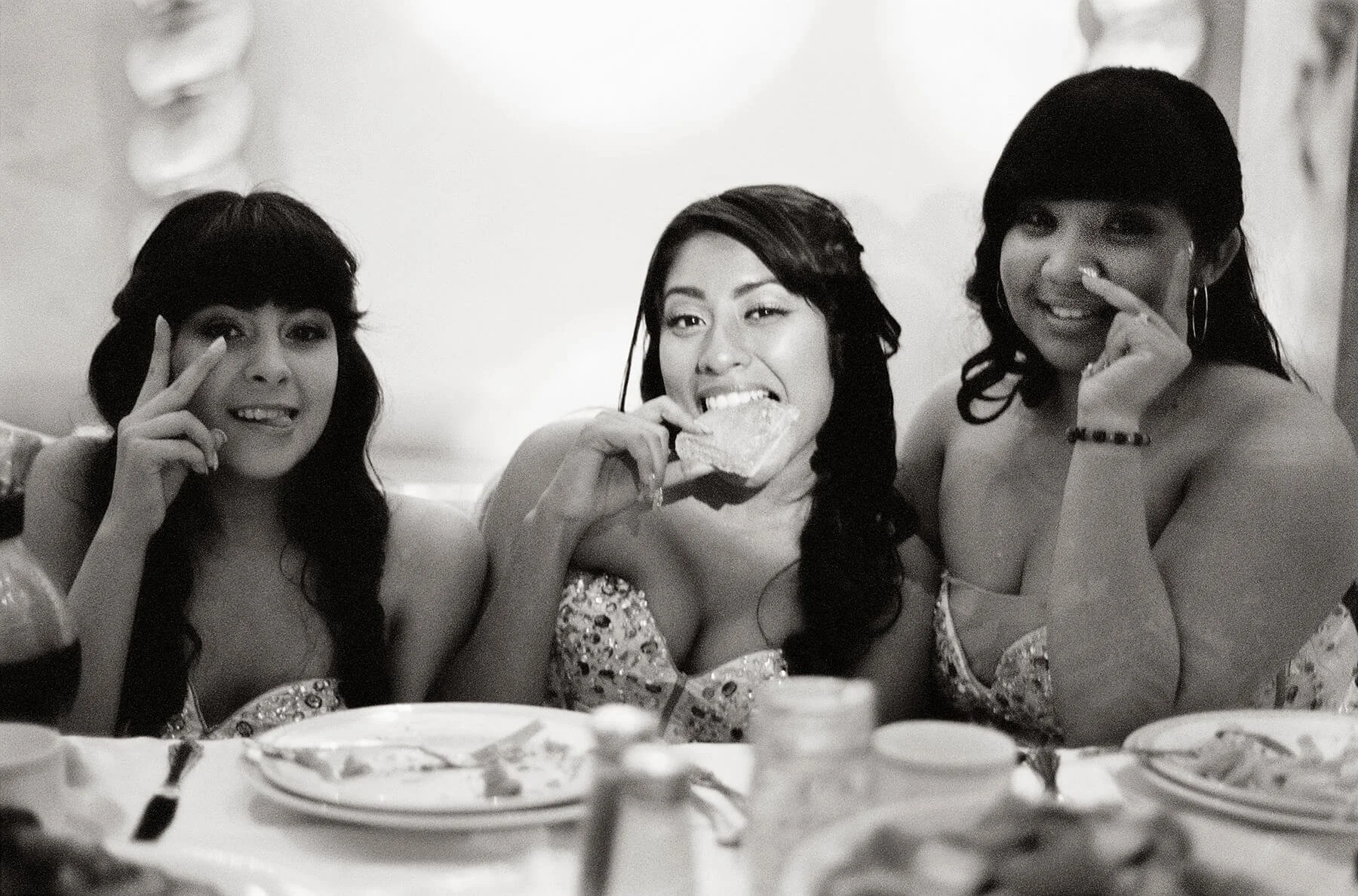 Three damas in party dresses sitting at a table, smiling, with one eating a slice of pizza.