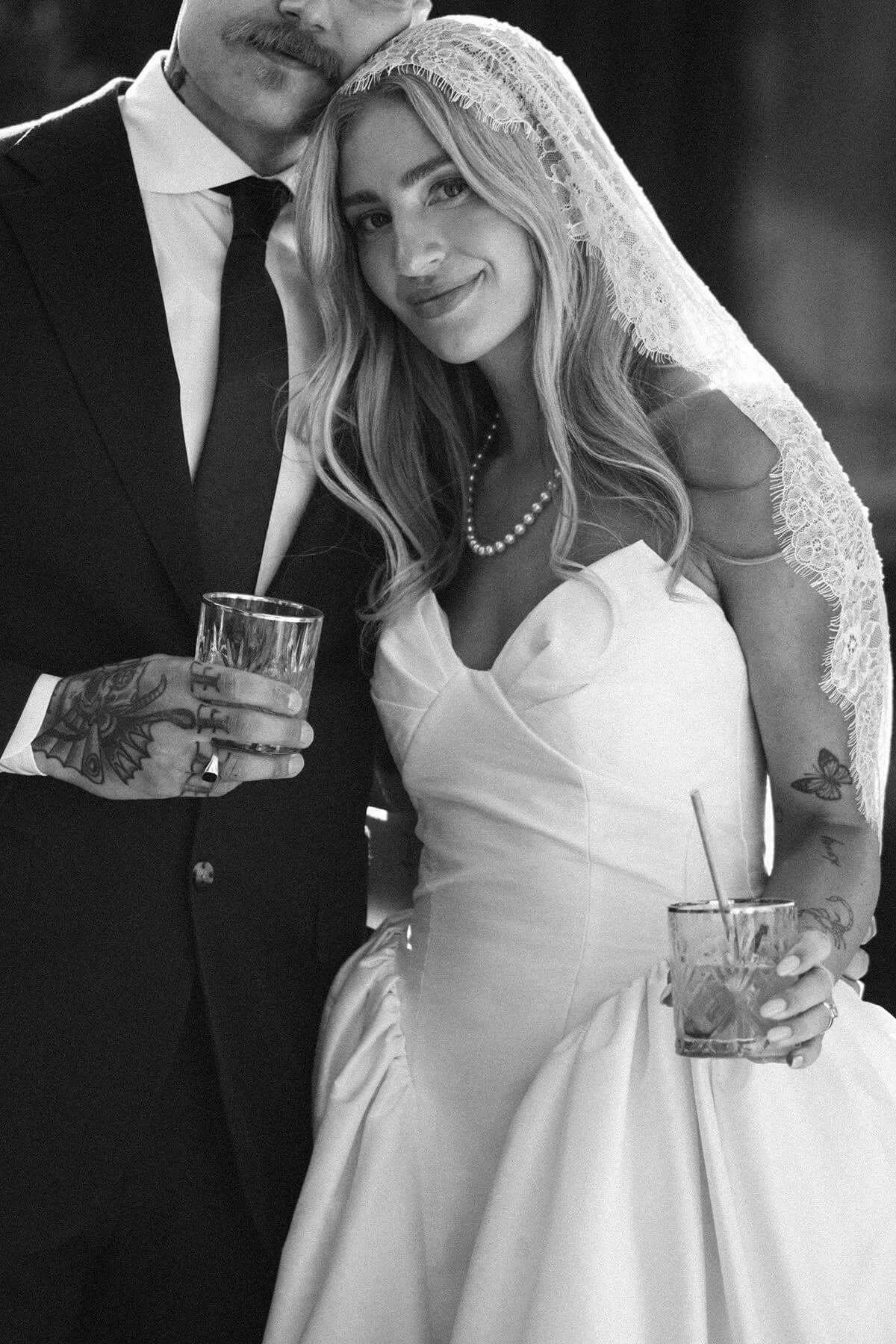 A black and white photo of a man in a tuxedo and a woman in a wedding dress, both holding drinks, standing together at a wedding celebration.
