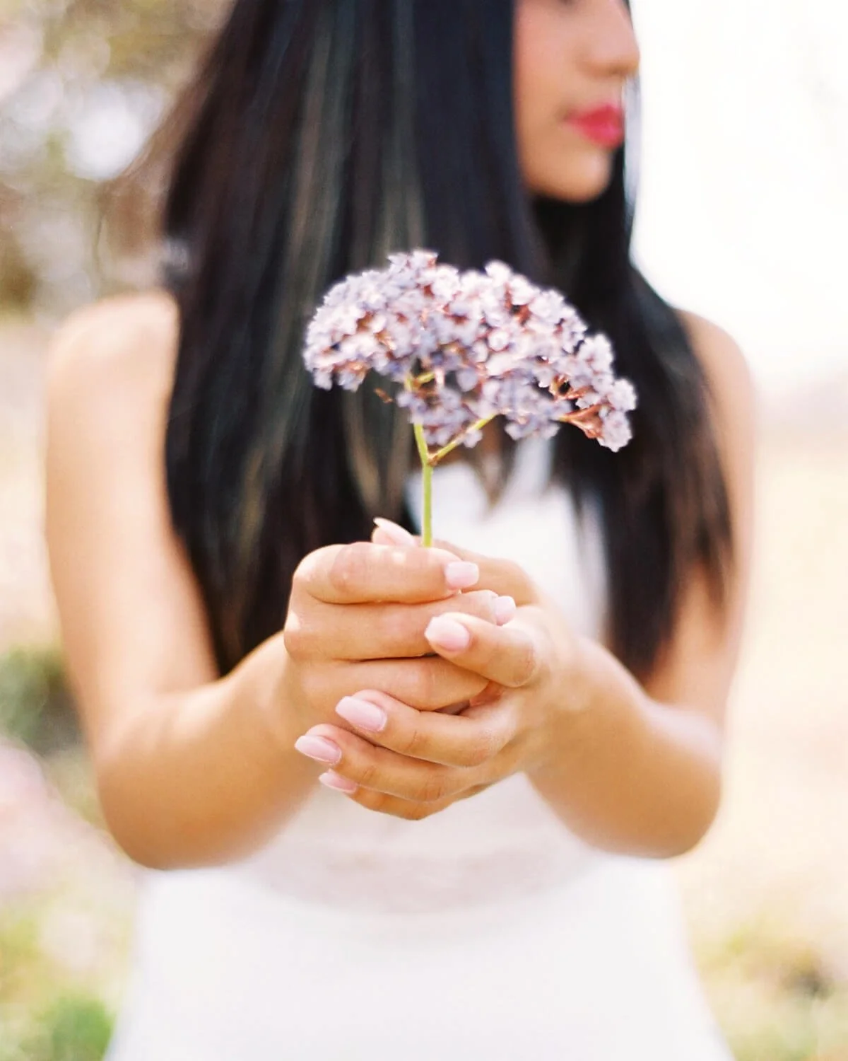A Quinceañera with long dark hair and pink lipstick holding a light purple flower with green stem, outdoors.