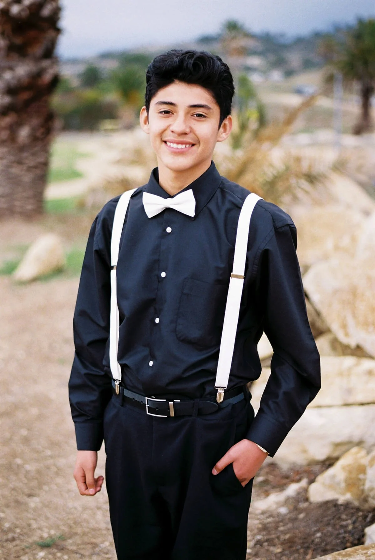 A chambelan in black dress shirt with white suspenders and bow tie standing outdoors on a trail with rocks and trees, smiling at the camera.