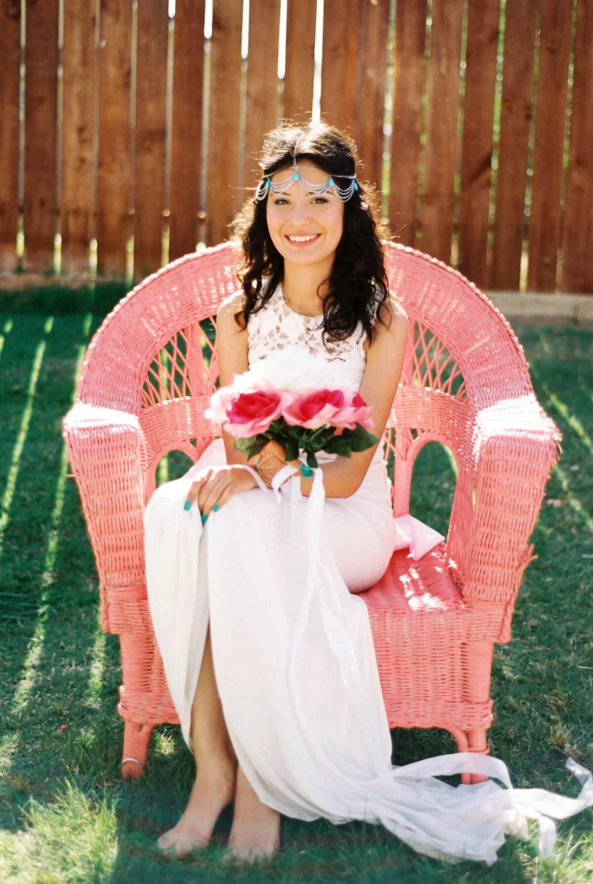 A Quinceañera in white dress sitting on pink wicker bench holding pink flowers, outdoors in backyard with wooden fence and green grass.