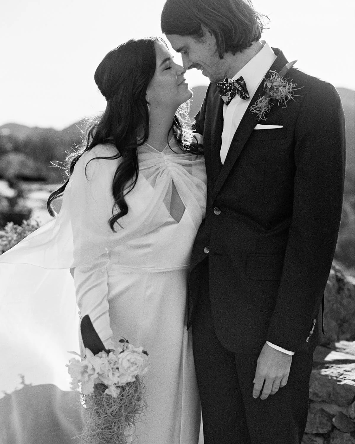 A black and white photo of a bride and groom on their wedding day in Cielo Farms, standing close with foreheads touching, smiling. The bride is holding a bouquet of flowers, and the groom is dressed in a tuxedo with a bow tie and a boutonniere.