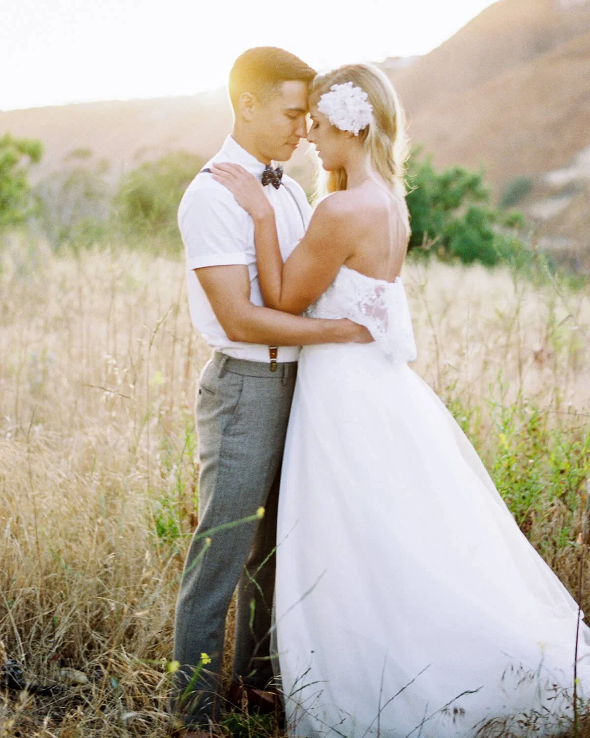 Newlywed couple embracing in a sunlit field, bride in a white wedding dress and groom in a white shirt and gray trousers.