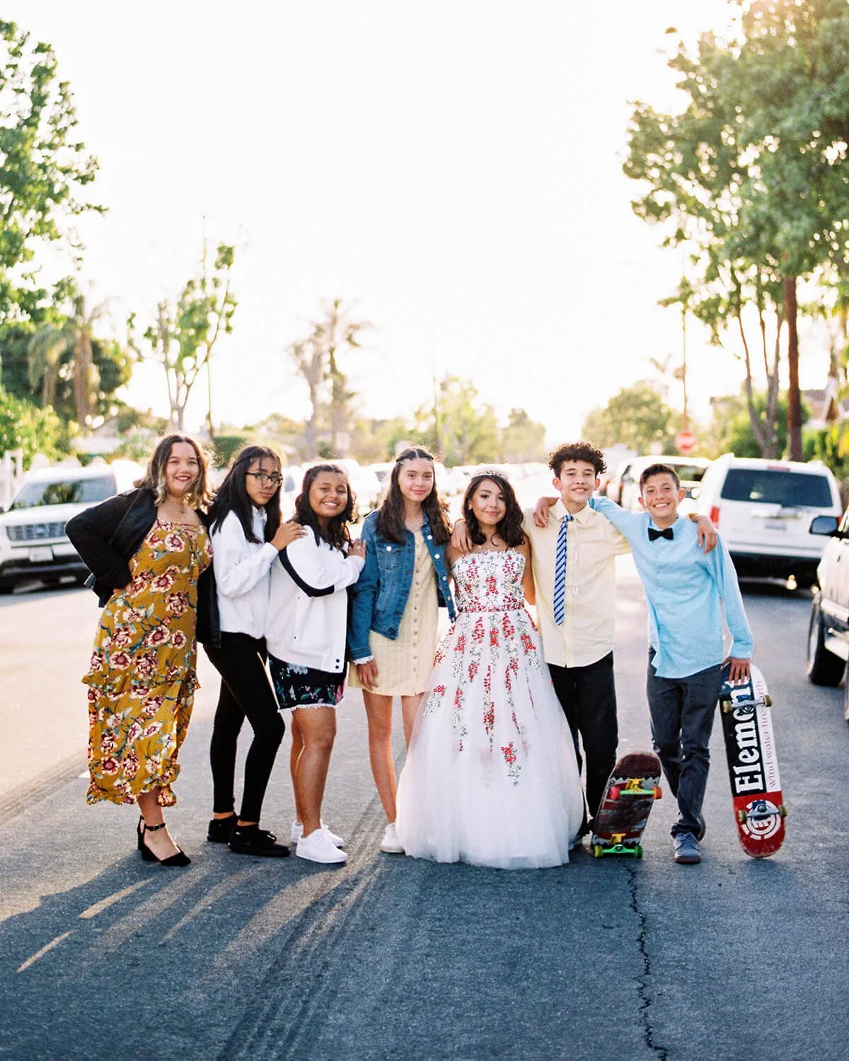 Group of seven diverse young people standing in a parking lot during daylight, with trees and parked cars, wearing casual and semi-formal clothing, some with skateboards, smiling at the camera with the Quinceañera in the middle of the group.