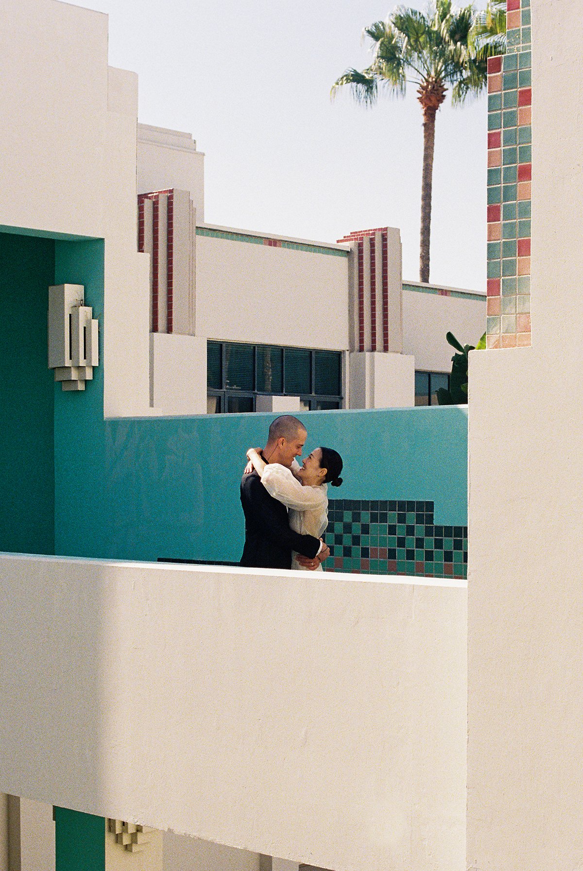 A couple dancing closely on a rooftop with modern architectural walls, a palm tree, and a clear sky in the background.