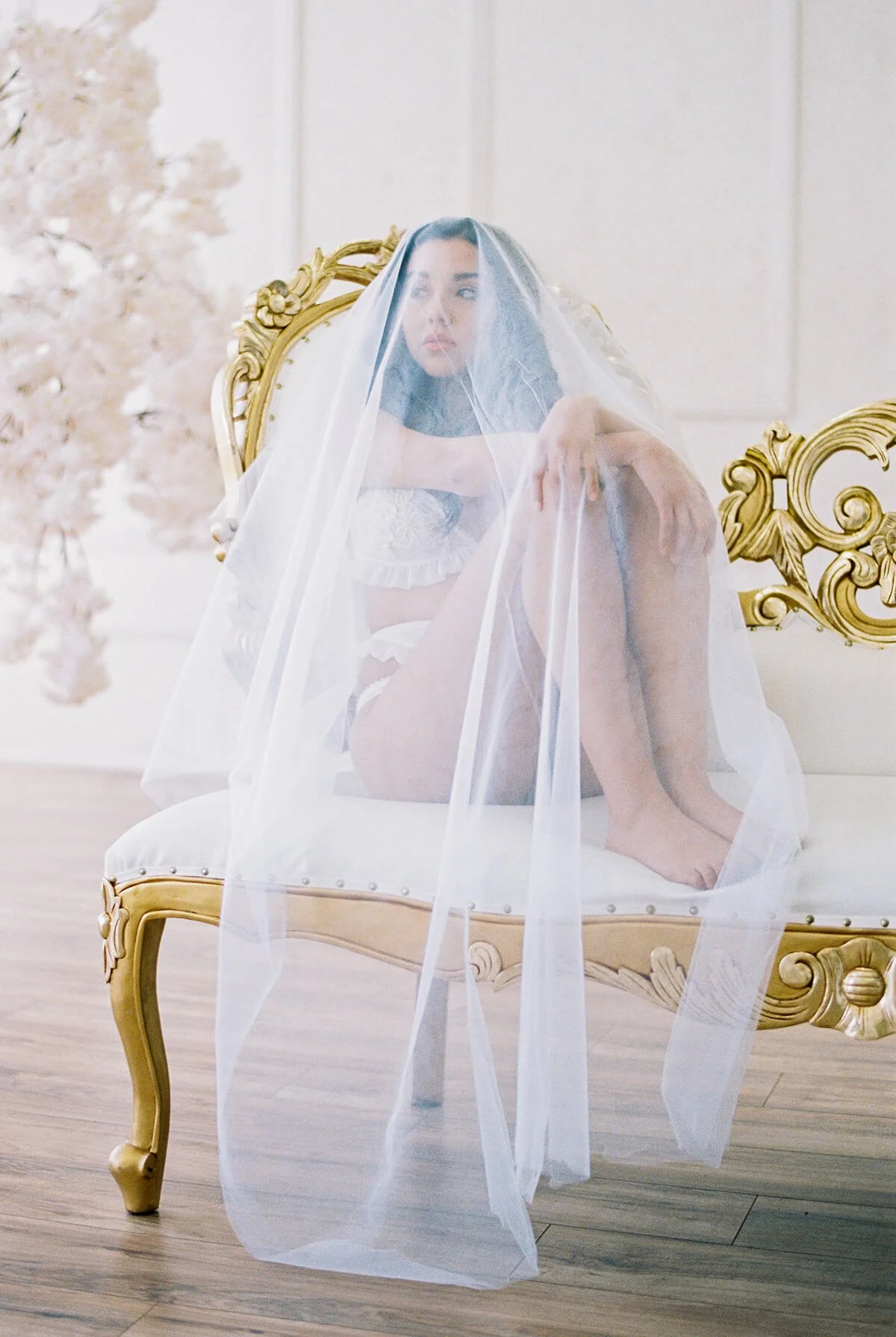A woman sitting on a gold ornate bench with a sheer veil over her head and shoulders, in a softly lit room with light-colored walls and wooden flooring.