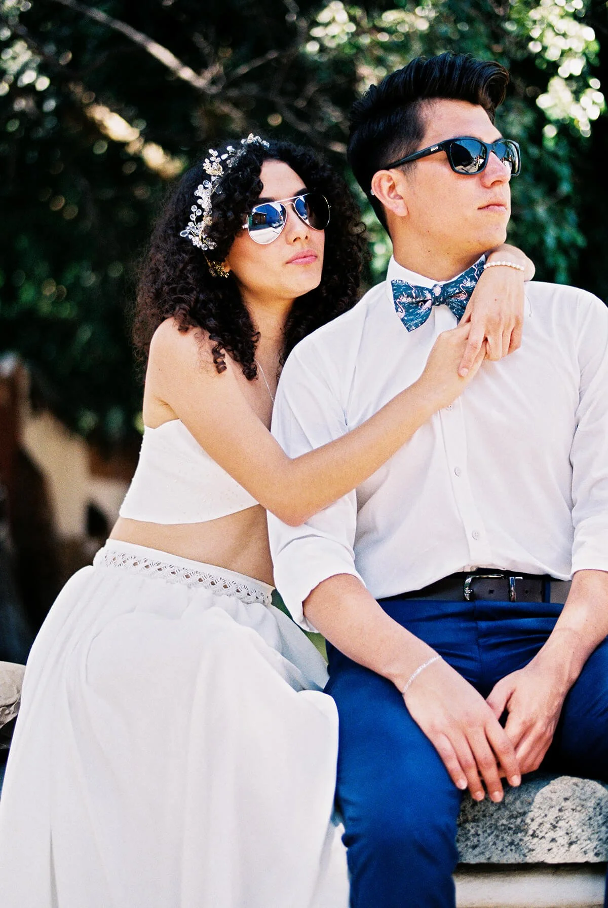 Couple wearing sunglasses poses outdoors, with the woman in a white dress and the man in a white shirt and blue bow tie.