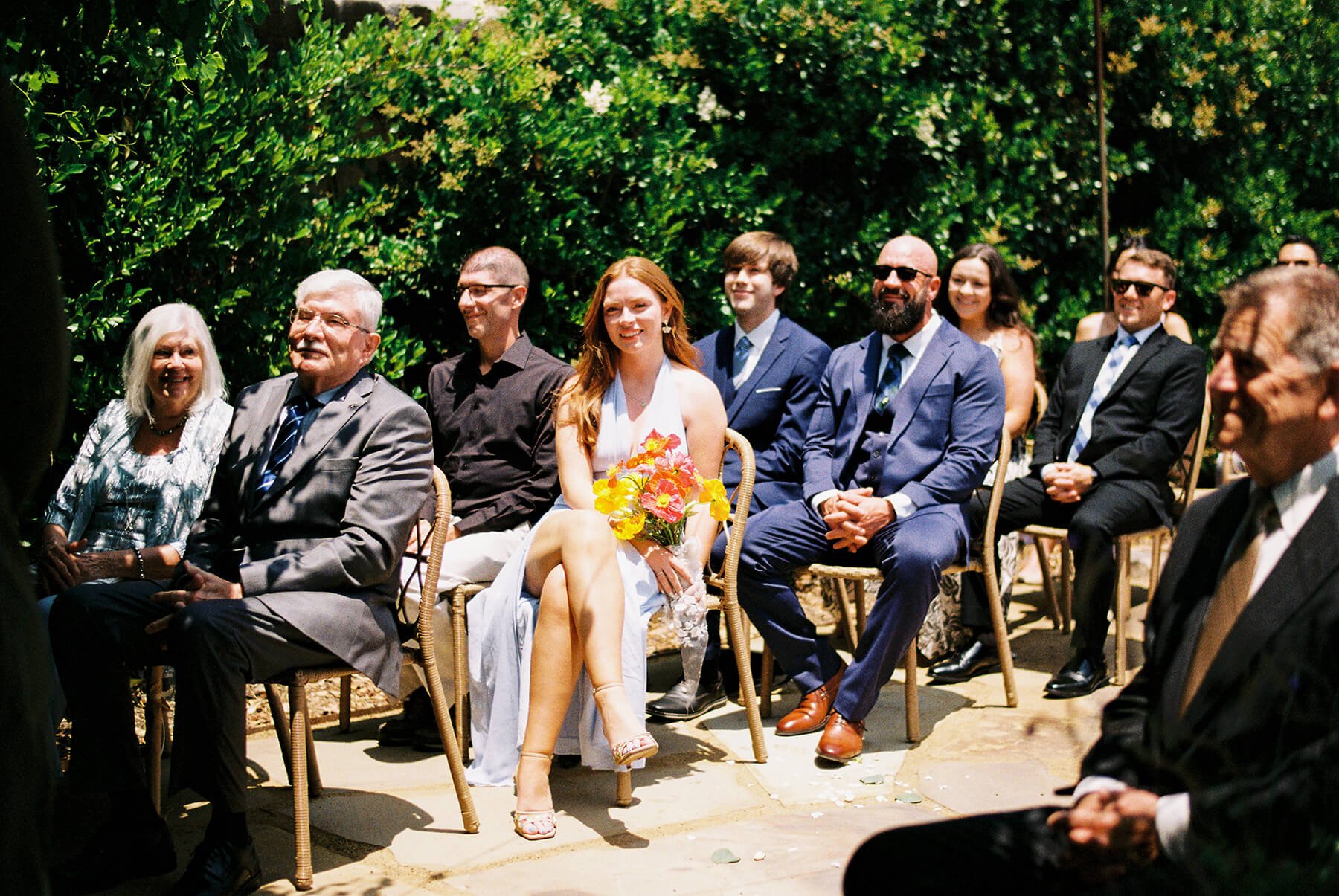 People attending an outdoor wedding ceremony, sitting on chairs, with the bride in a white dress holding a bouquet of flowers, and guests dressed in formal attire, surrounded by green foliage.