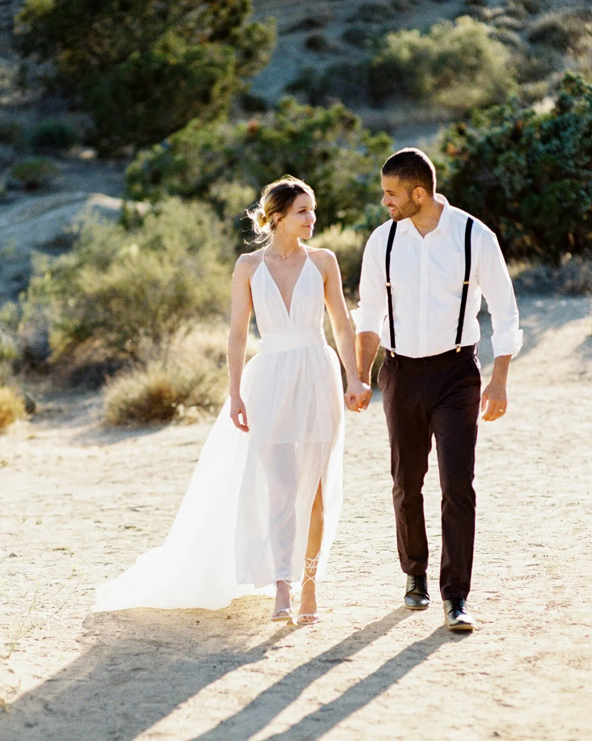 A bride and groom walking hand in hand outdoors on a sunny day, surrounded by desert vegetation in Griffith Park.