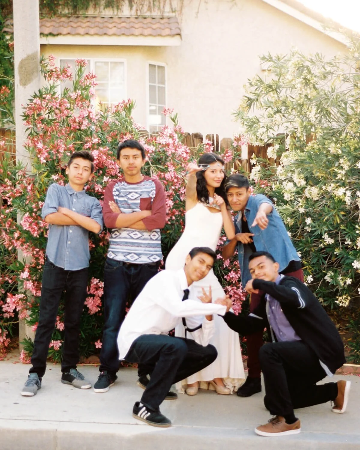 Group of six teenage friends posing outdoors in front of flowering bushes and a house, some making gestures and smiling.