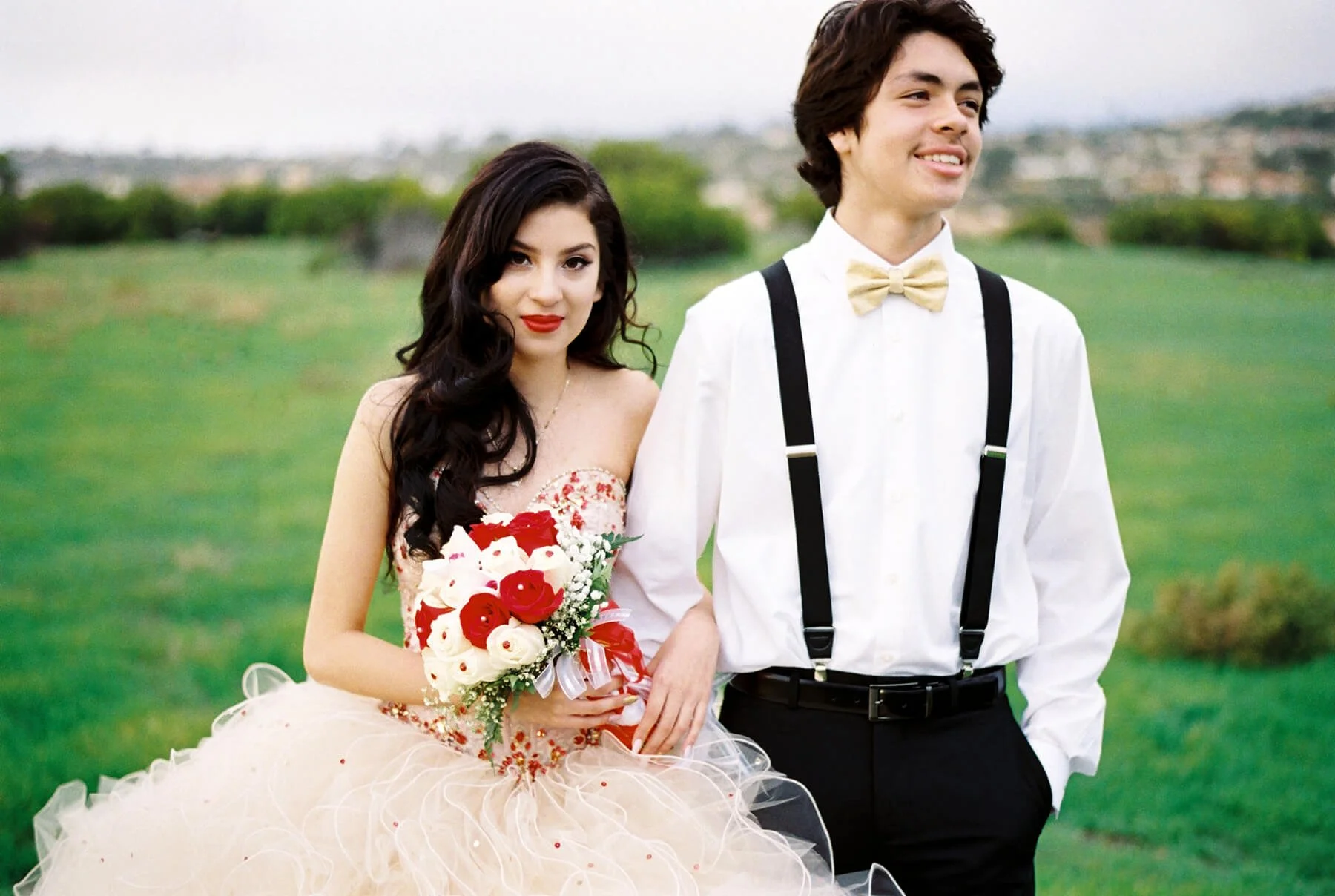 A Quinceañera holding a bouquet of white and red flowers, standing next to the chambelan de honor in a white shirt, yellow bow tie, and black suspenders in an outdoor green field.