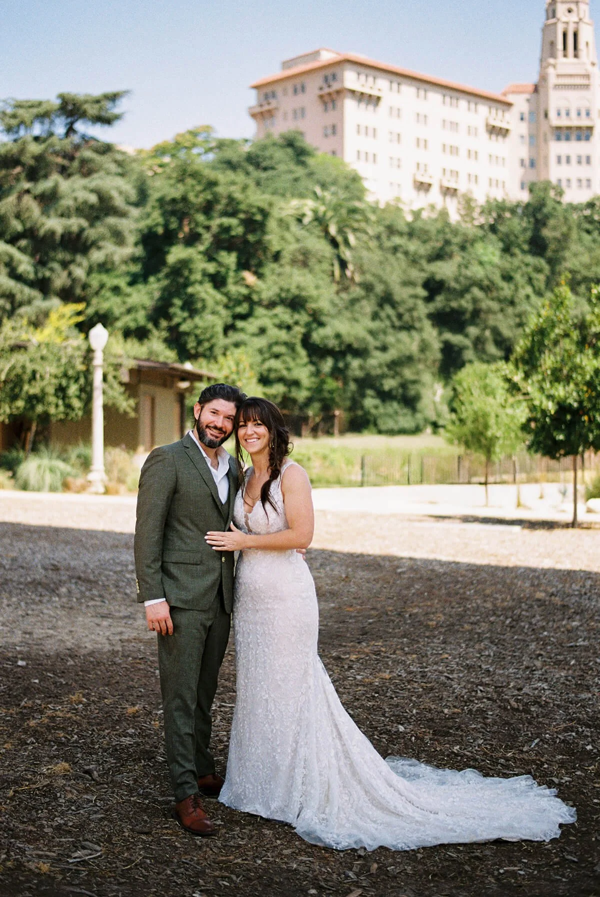 A bride and groom standing outdoors on a dirt path, smiling and embracing, with trees and a building in the background.