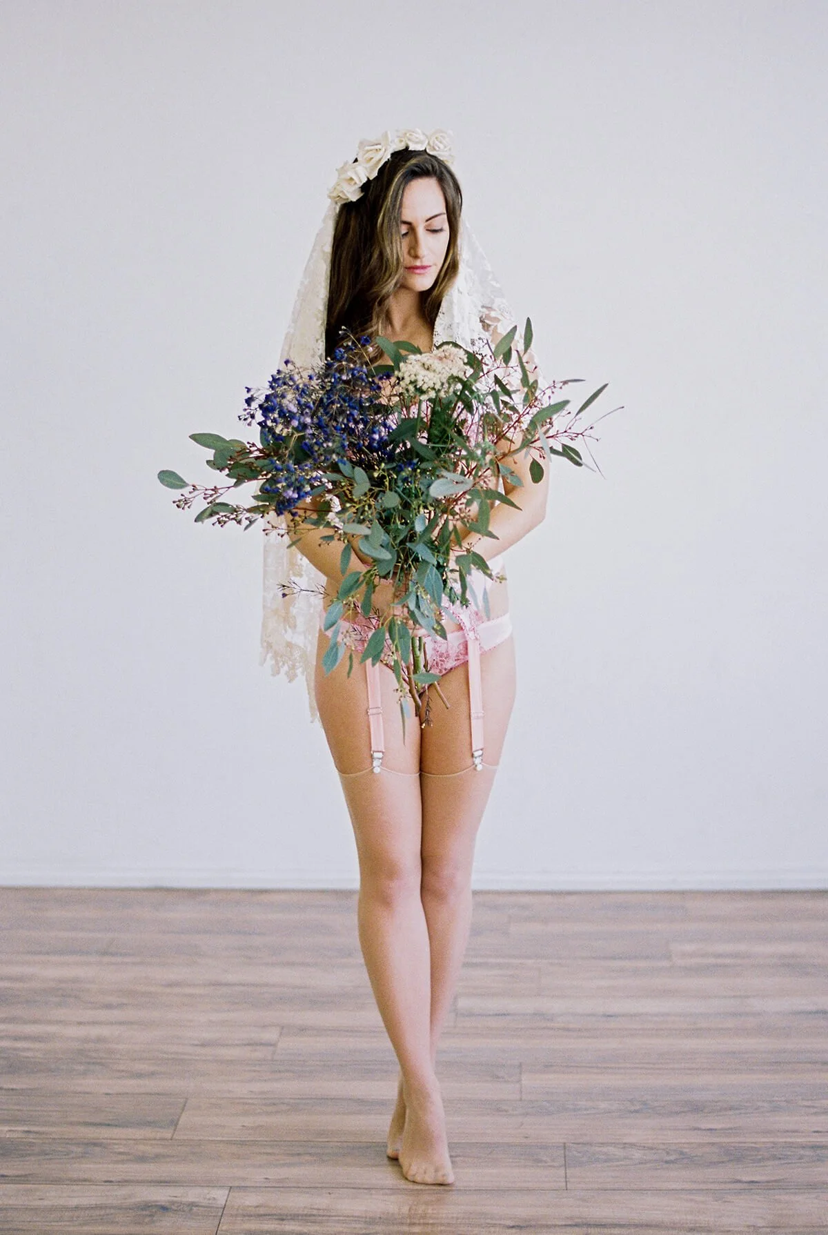 Bride wearing a flower crown and veil, holding a large colorful bouquet in a bridal lingerie photoshoot