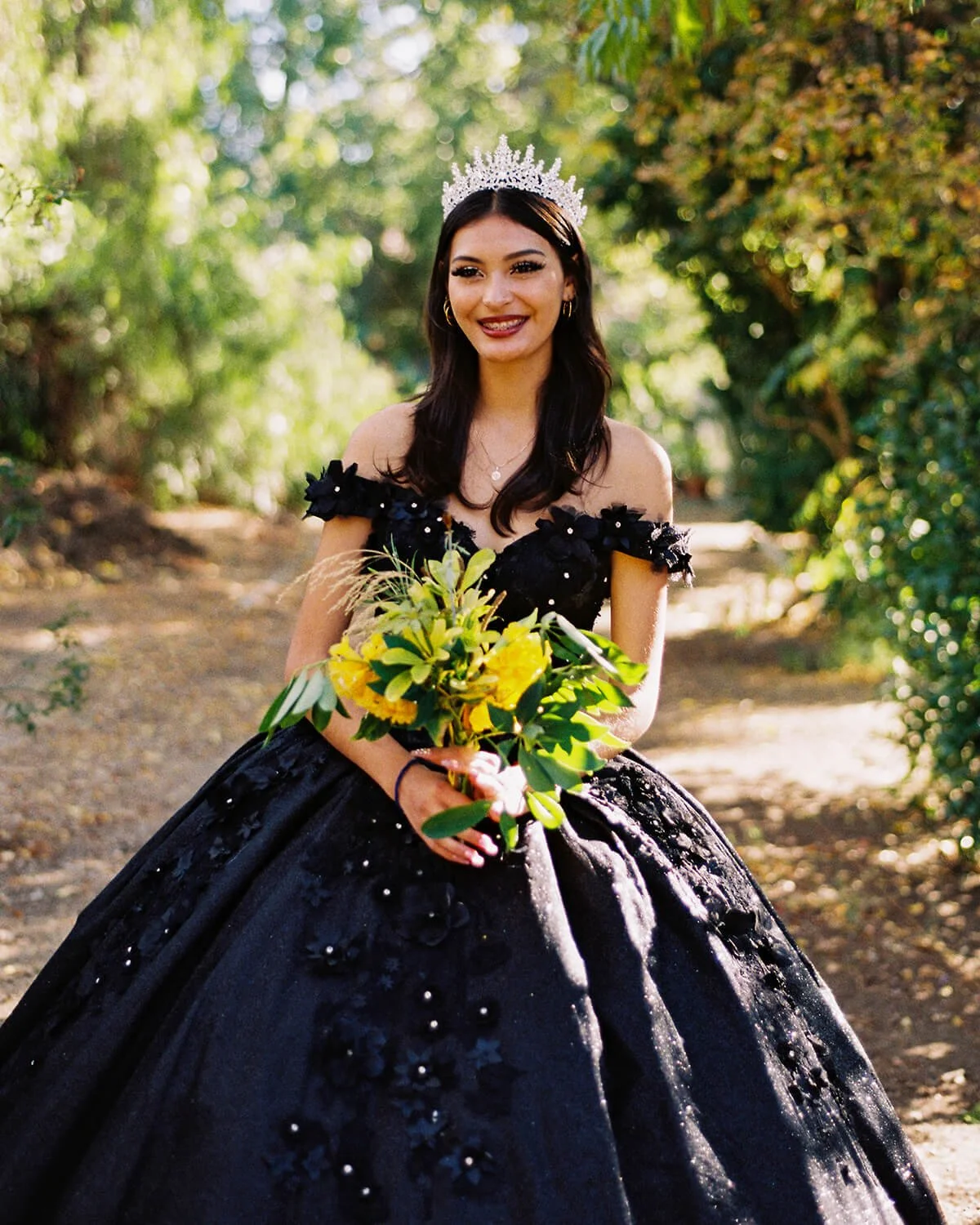 A Quinceañera in a black gown with floral details and a tiara, holding a bouquet of yellow flowers, standing outdoors on a sunny day.