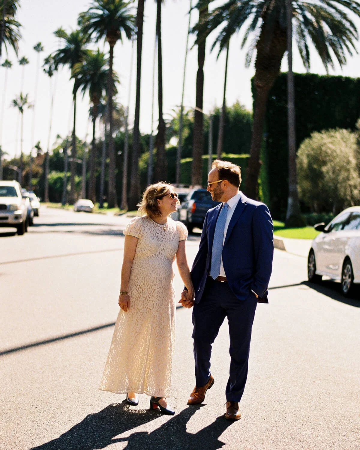 A woman in a cream lace dress and a man in a navy suit holding hands and smiling at each other on a sunny street lined with tall palm trees.