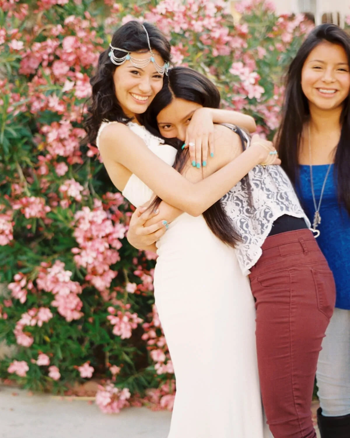 Three women embrace in front of pink flowering bushes, smiling and happy.