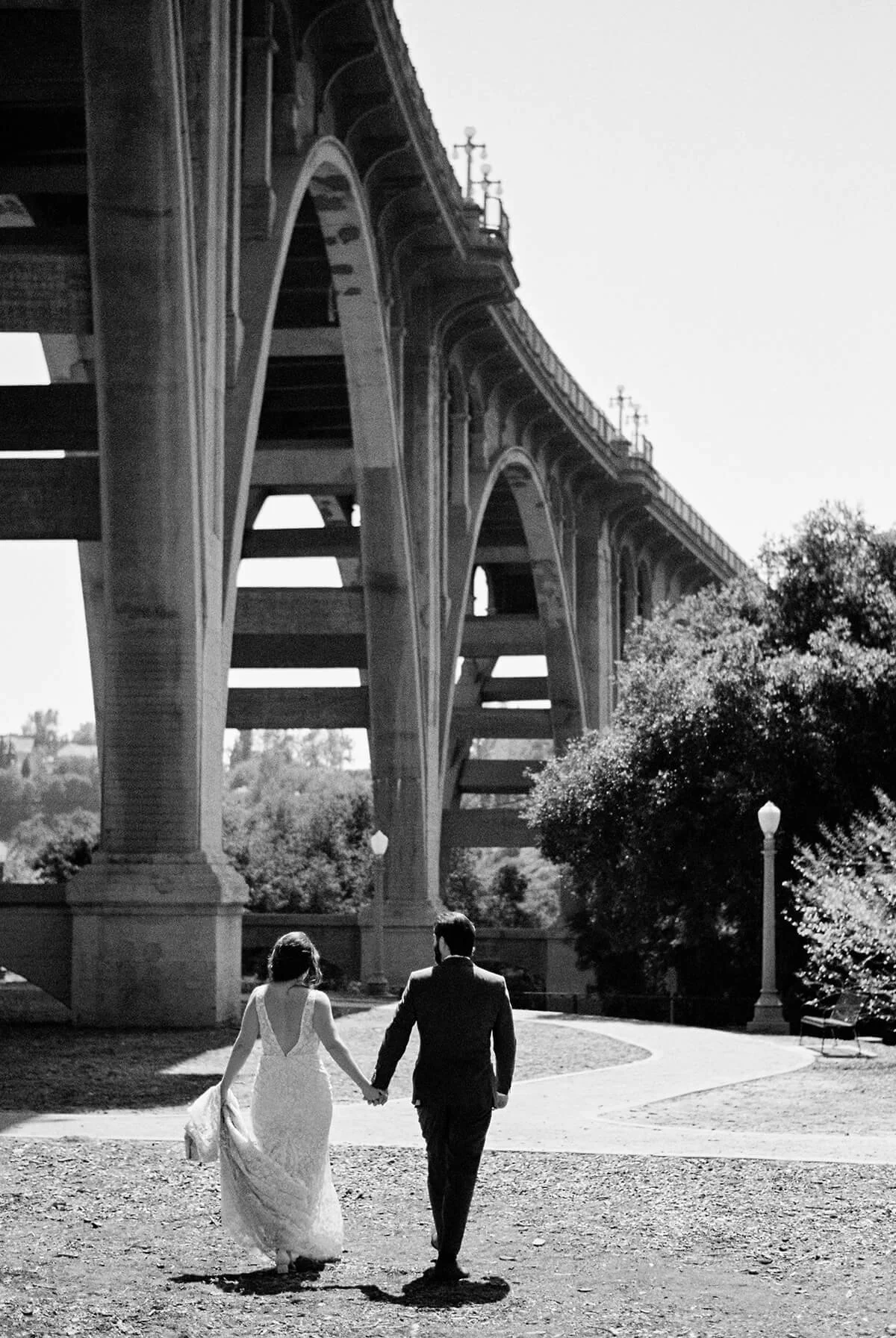 A black-and-white photo of a couple in wedding attire holding hands while walking under a large bridge.