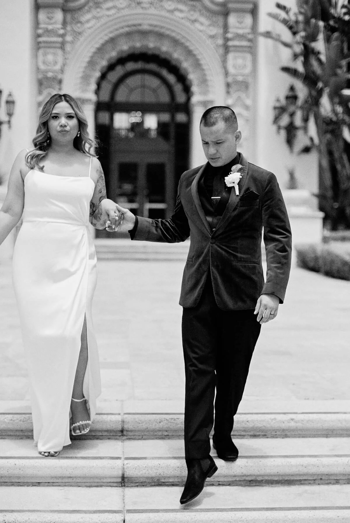 A black and white photo of a couple, with the woman in a white dress and the man in a tuxedo, walking down stairs holding hands outside a building with ornate architecture.