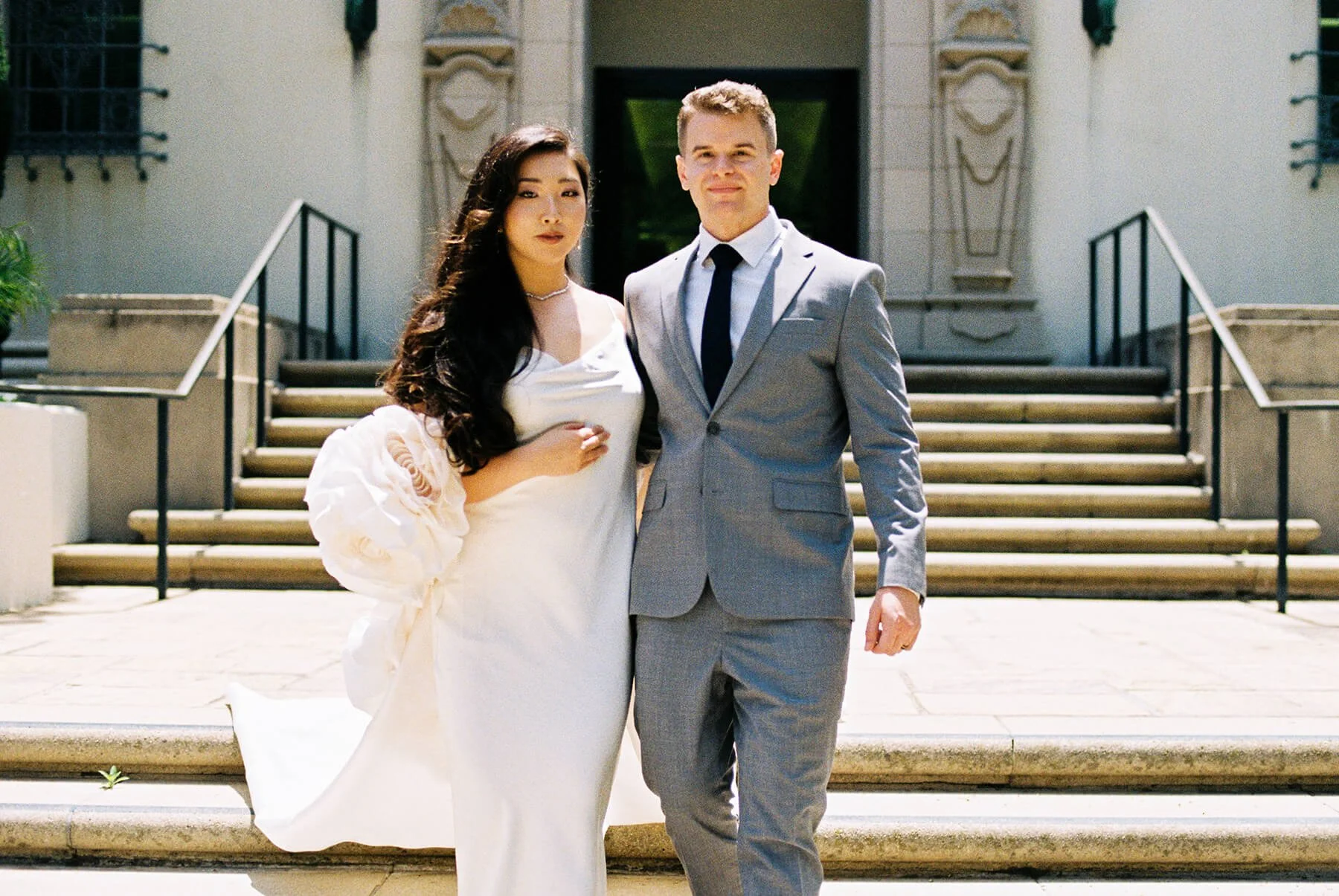 A newlywed couple standing side by side on the steps of a building. The woman wears a white wedding gown and has dark, wavy hair. The man wears a light gray suit with a white shirt and a dark tie. They are outdoors on a sunny day.