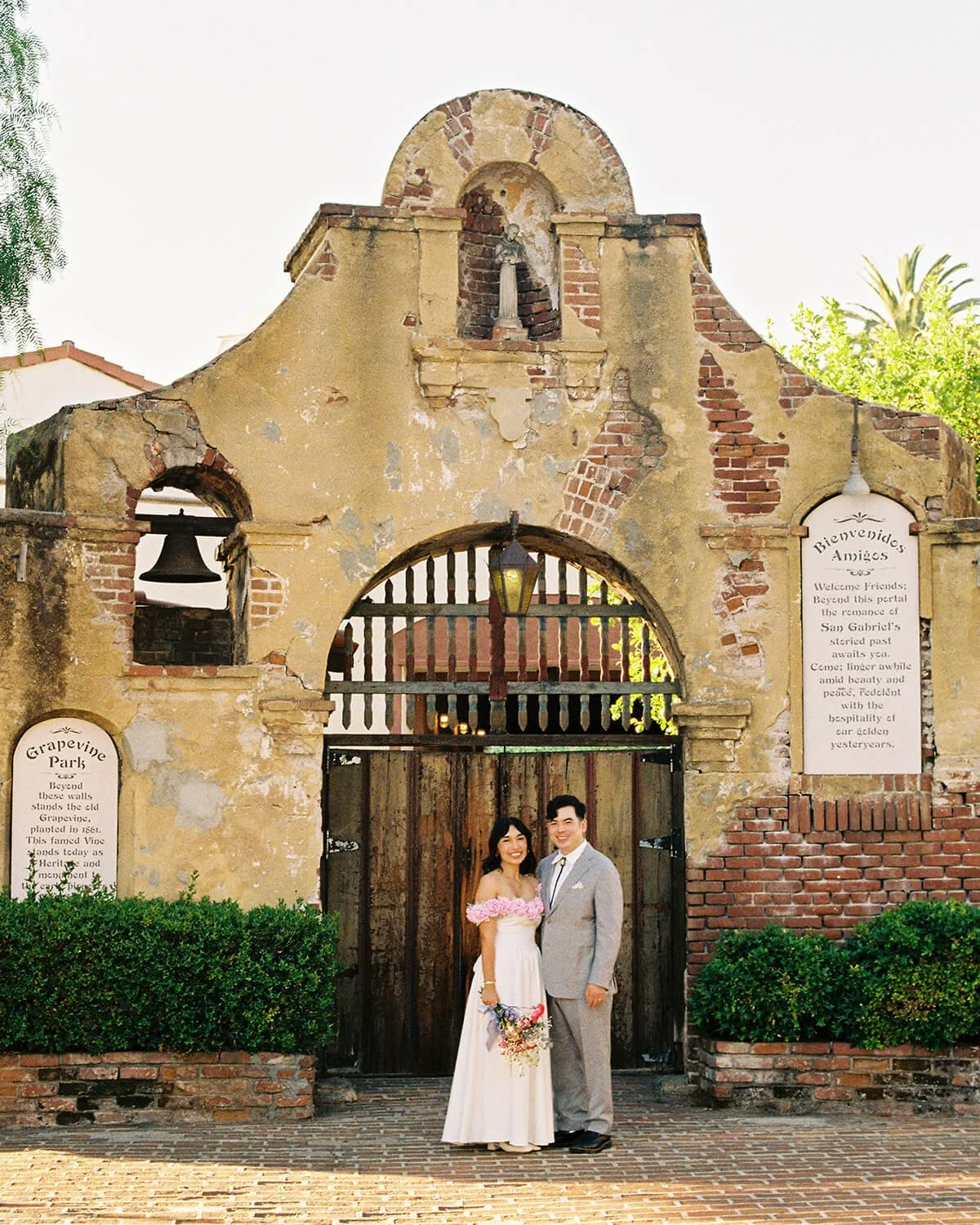 A newly married couple standing in front of the old brick and stone archway gate of the Grapevine Arbor in San Gabriel, with welcoming signs on either side, greenery at the base, and a clear sky above.