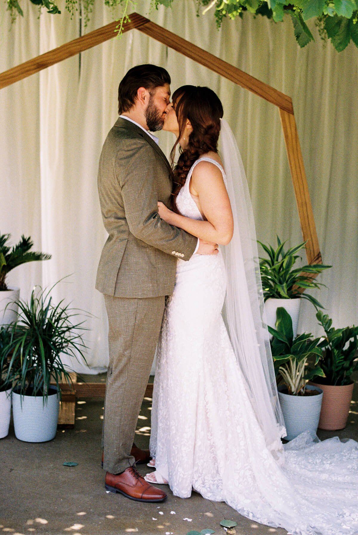 A bride and groom share a kiss during their wedding ceremony, standing under a wooden archway surrounded by green plants.