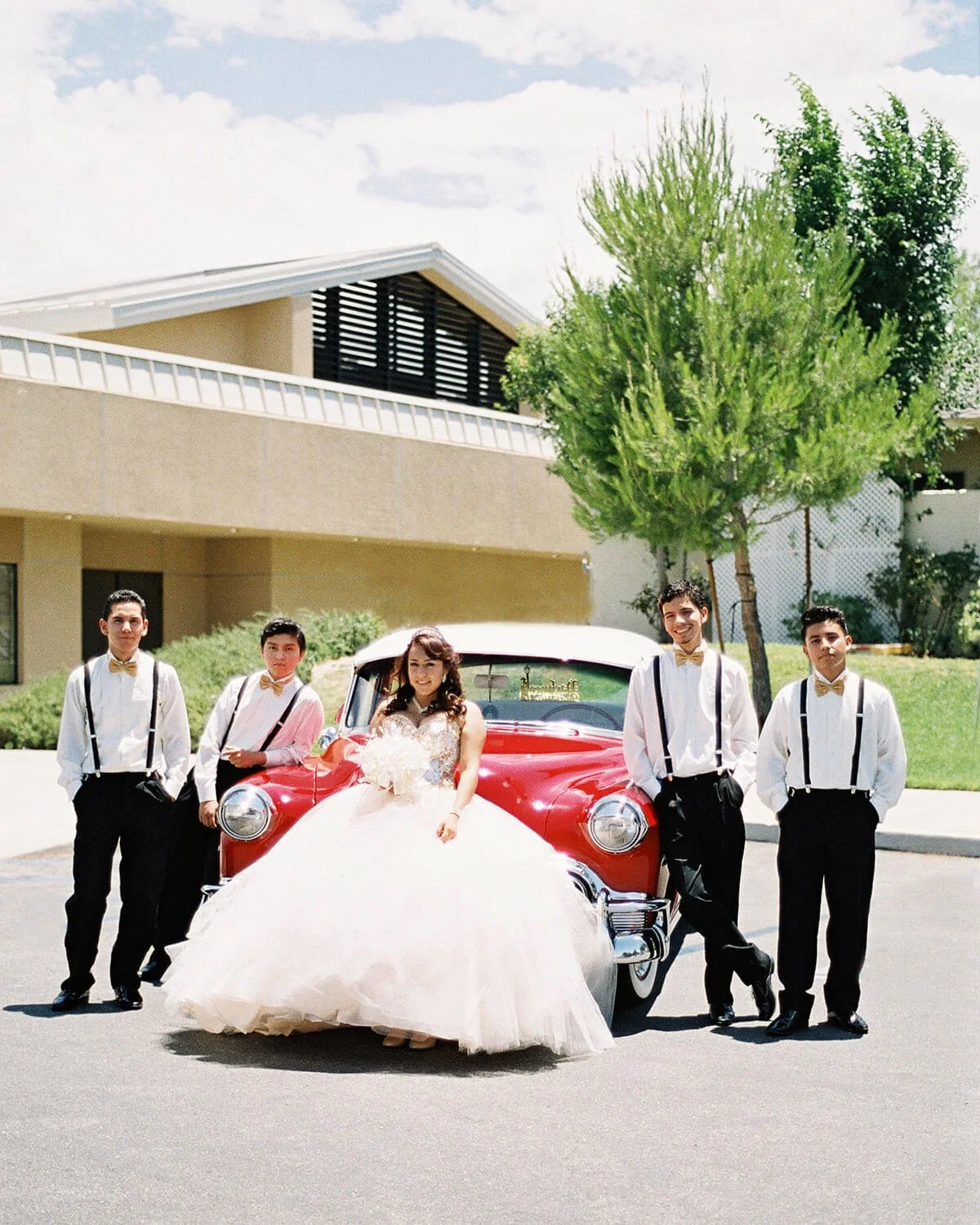 A Quinceañera in a white wedding dress sitting on the front bumper of a red vintage car, with five chambelanes in white shirts, black suspenders, and bow ties standing beside her.