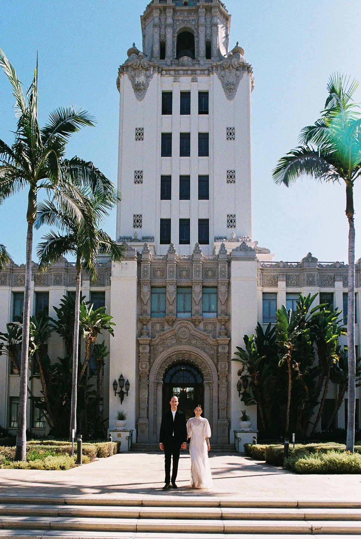 A couple in wedding attire walking hand in hand in front of an ornate historic hotel entrance with a tall tower and palm trees.