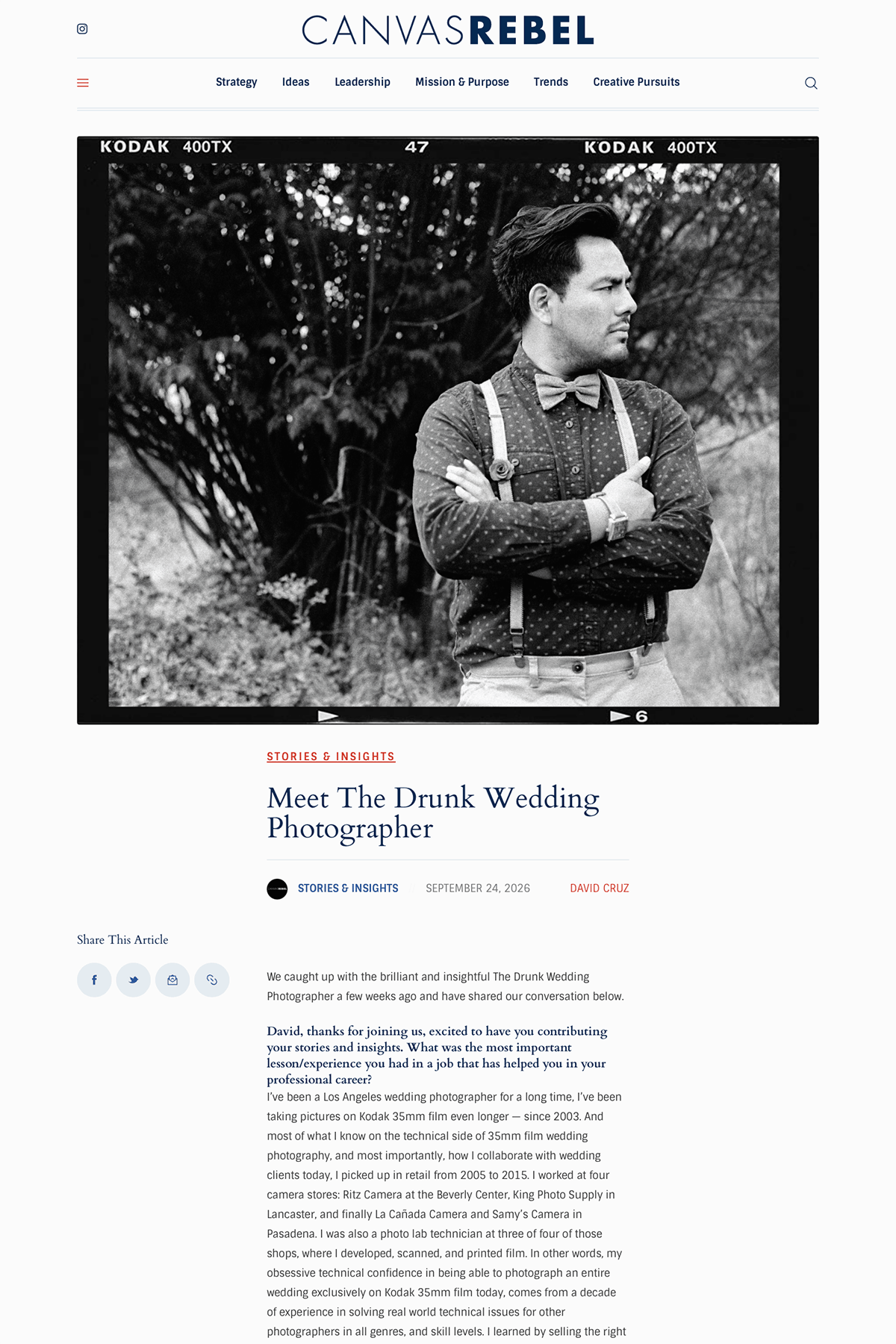 Black-and-white photo of a The Drunk Wedding Photographer in a polka dot shirt, suspenders, and a bow tie, standing outdoors with arms crossed, in front of trees.