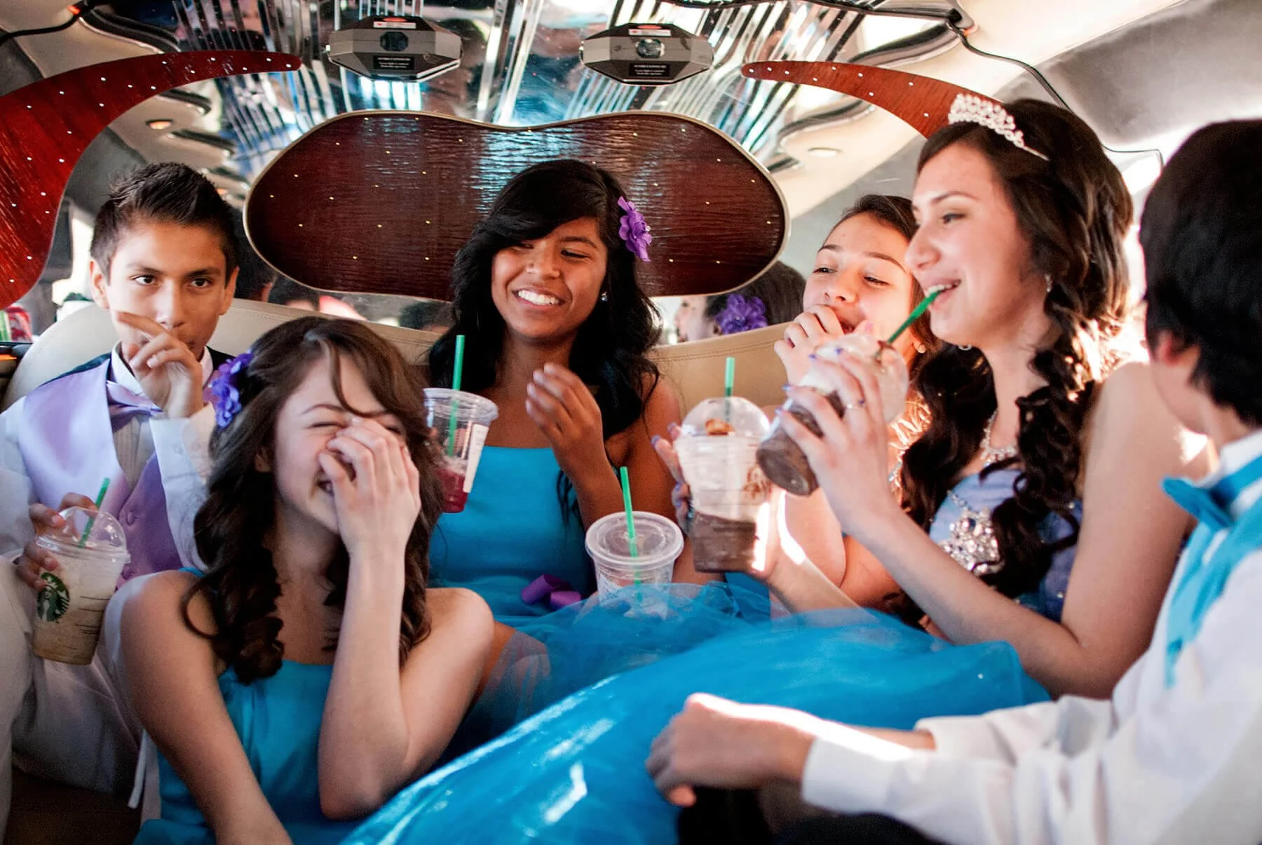 A Quinceañera Court dressed in formal and fancy attire, enjoying Starbucks drinks and laughing together inside a limousine with reflective ceiling panels.