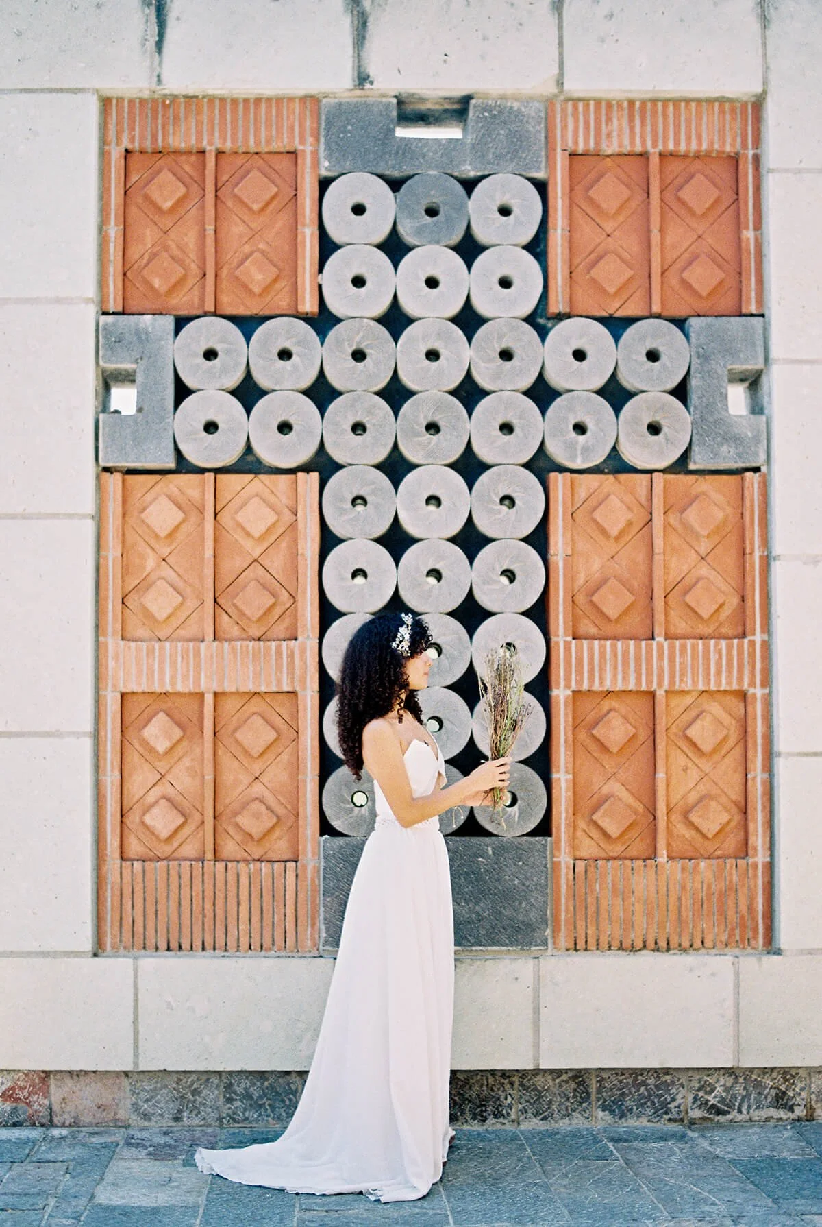 A woman in a white dress holding a bouquet of dried flowers, standing in profile against an ornate brick and stone wall with a decorative cross design. Cruz de Piedra in Oaxaca.