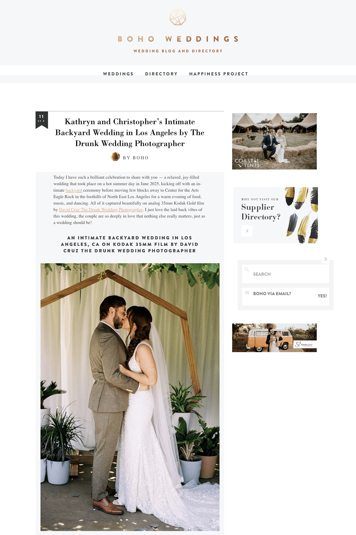 A wedding photograph showing a bride and groom standing close together, holding each other, under a wooden arch with green foliage. The bride is wearing a white lace wedding gown and veil, and the groom is in a gray suit. They are outdoors, surrounde