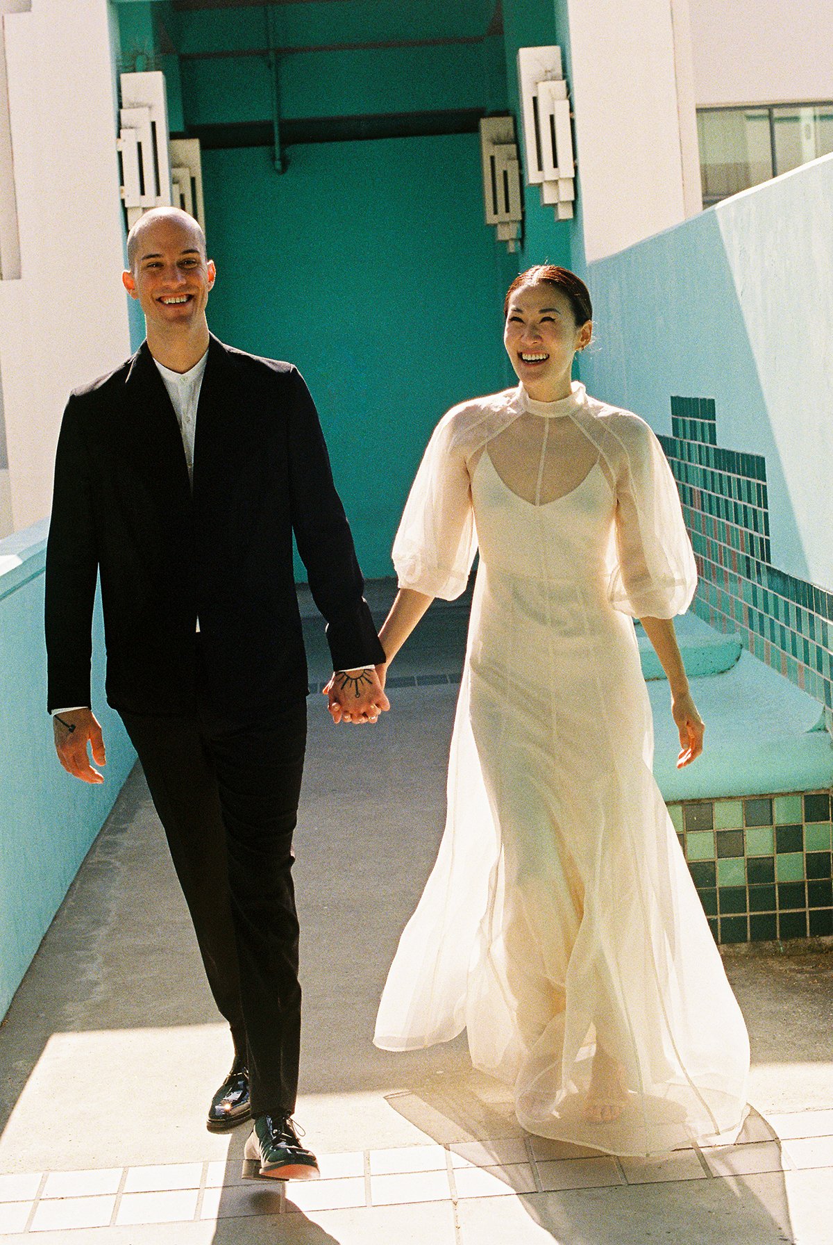 A smiling couple holding hands and walking outdoors, dressed in wedding attire—man in a black suit and woman in a white dress, on a sunny day near a building with teal and white walls.