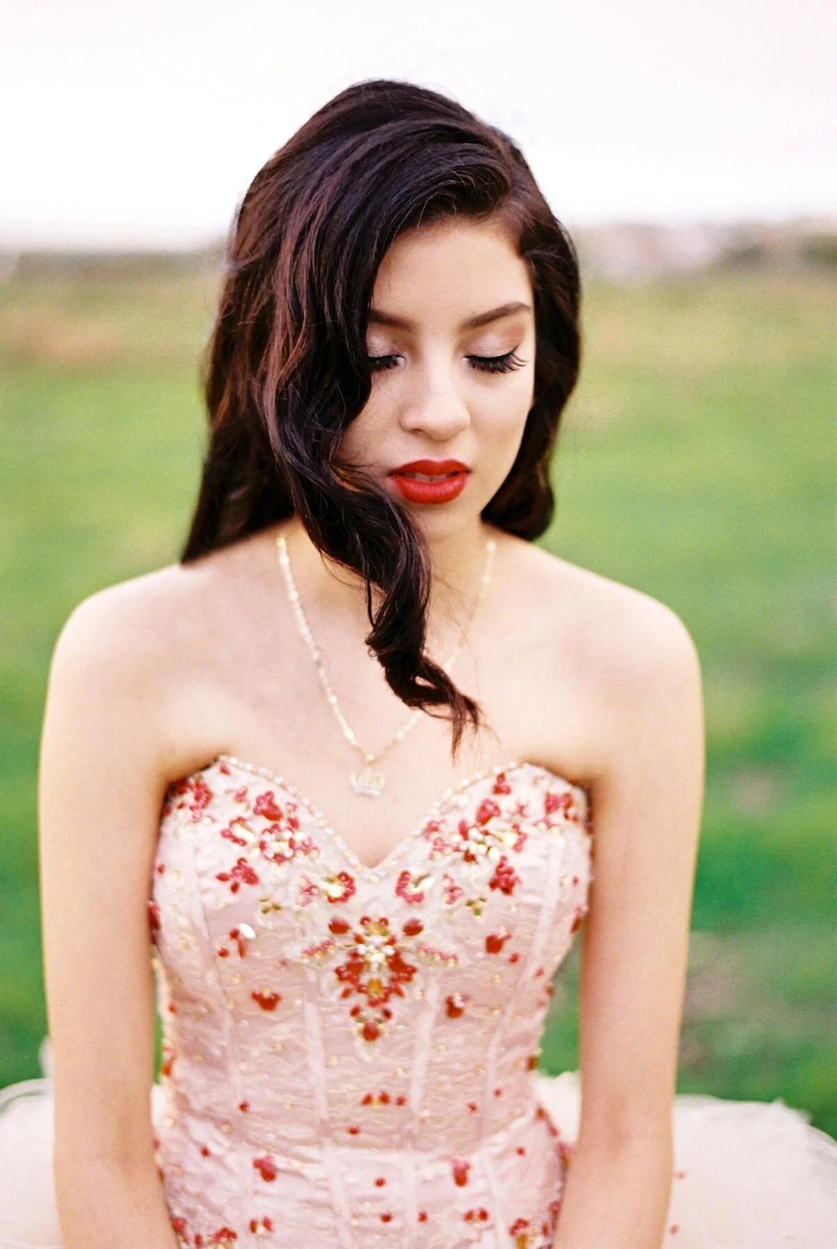 A Quinceañera with dark, wavy hair and red lipstick wears a strapless pink dress with floral embroidery and a pearl necklace, standing outdoors with a blurred green background.