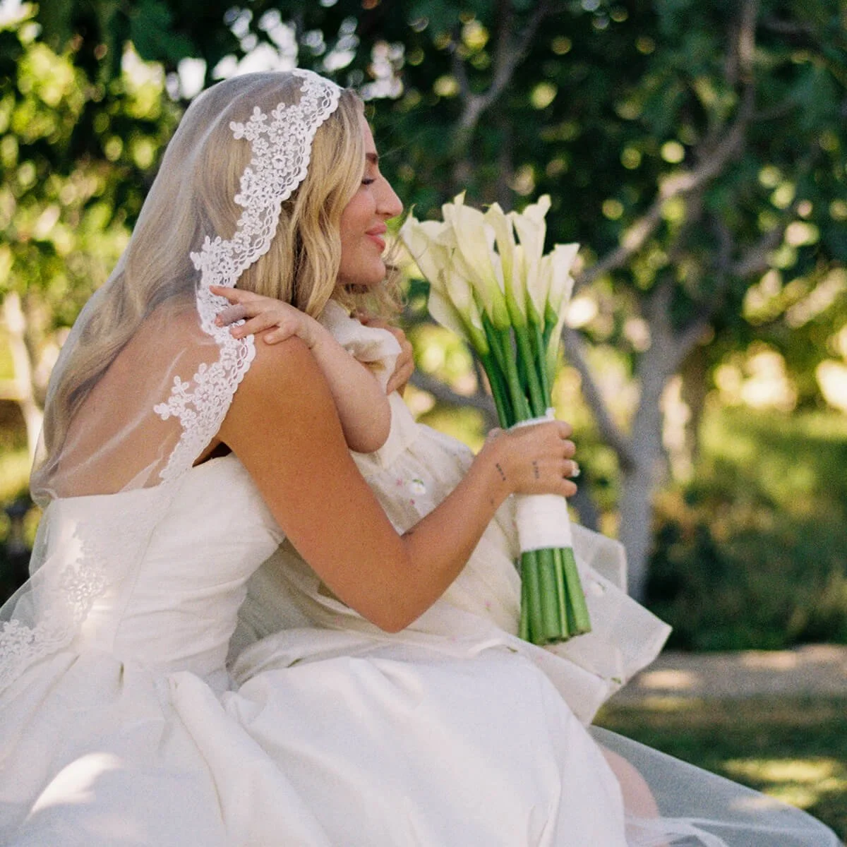A bride in a white wedding dress with a lace veil holding a bouquet of white calla lilies, smiling with her eyes closed outdoors surrounded by greenery.