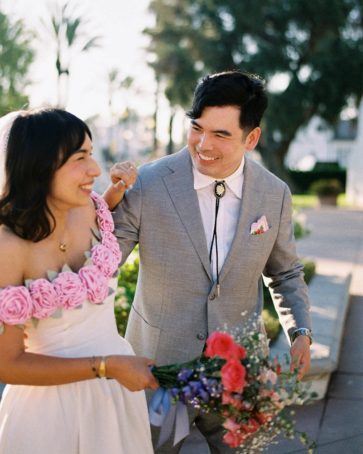 A couple exchanging smiles during their wedding, the bride holding a bouquet of pink and purple flowers, outdoors with trees and sunlight behind them.