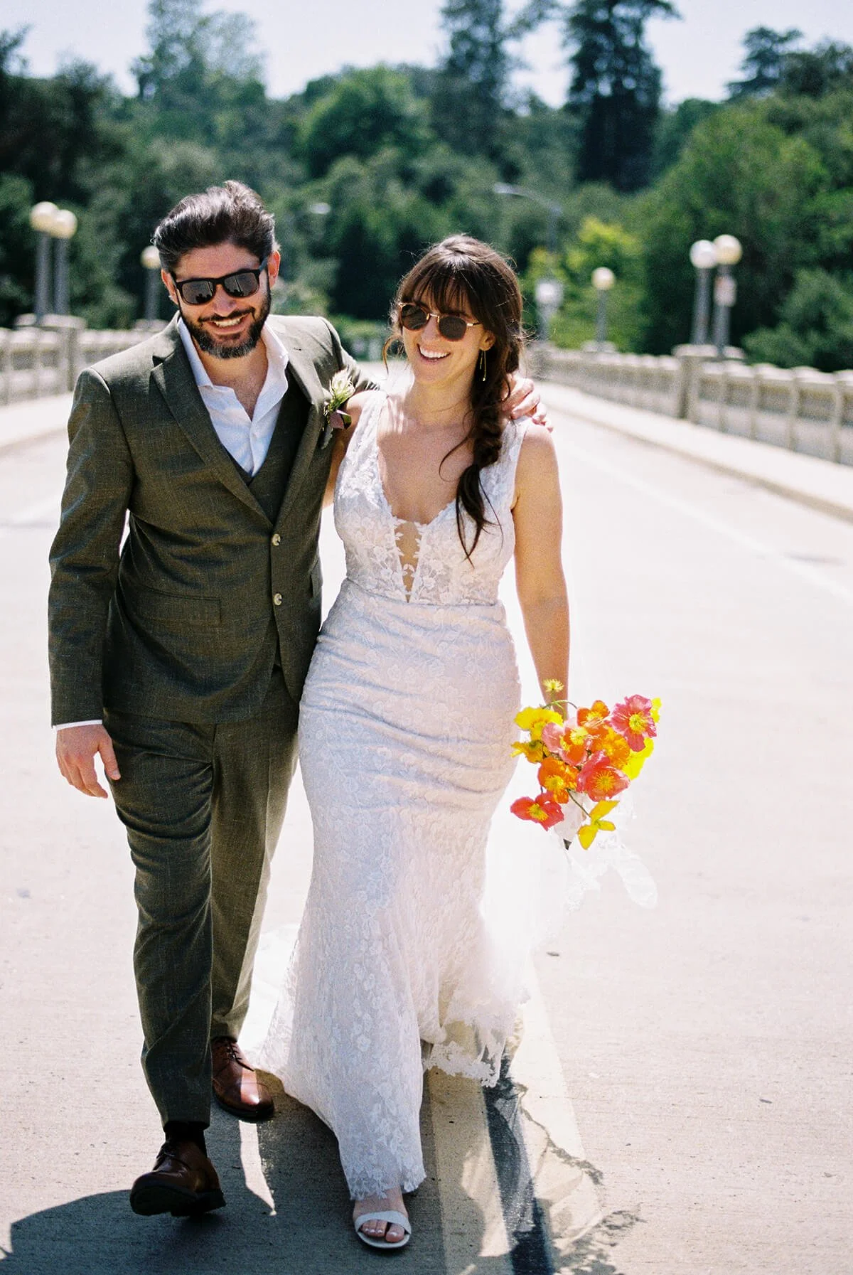 A newlywed couple walking arm-in-arm on a sunny outdoor bridge, smiling. The bride is wearing a lace white wedding gown, holding a colorful bouquet, and wearing sunglasses. The groom is wearing a dark suit with a white shirt, sunglasses, and has a bo