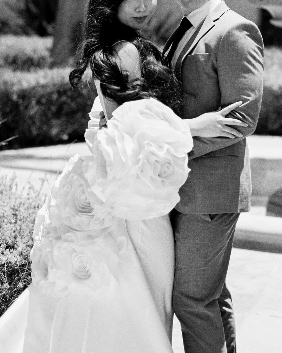 Black and white photo of a couple embracing outdoors, with the woman wearing a dress with floral details and the man in a suit.