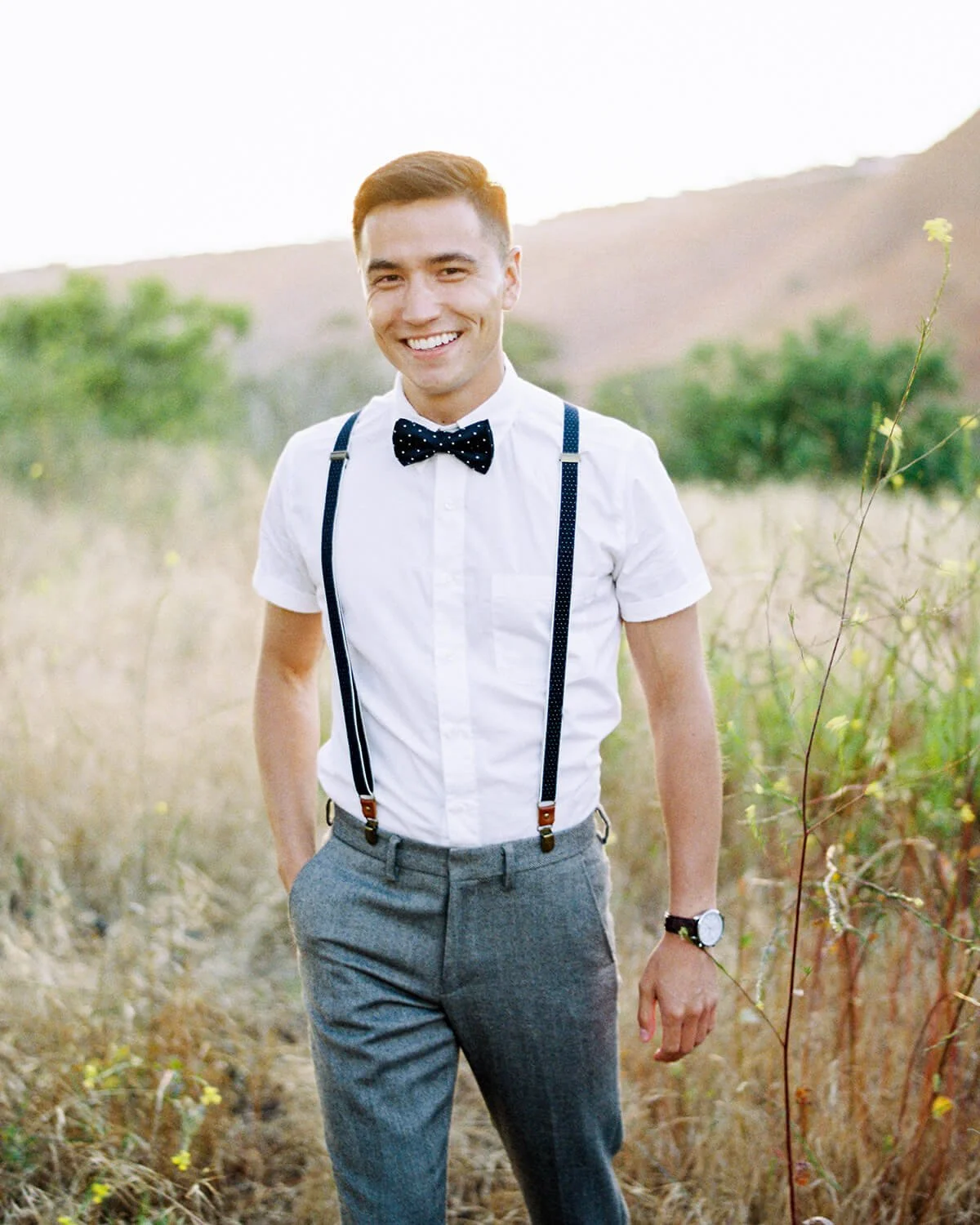 A young man smiling outdoors wearing a white short-sleeve shirt, a black bow tie with white dots, gray pants, suspenders, and a watch, standing in a grassy field with hills and greenery in the background.