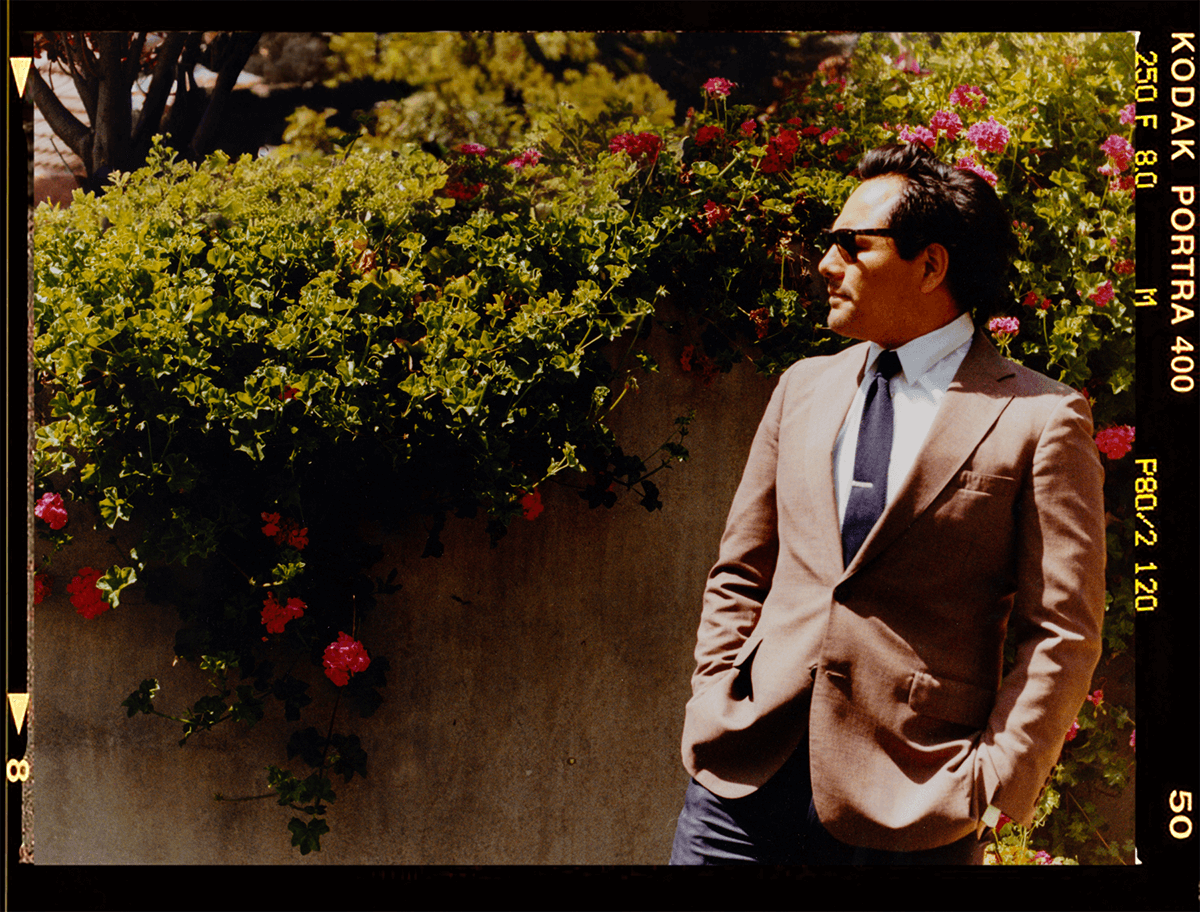 David Cruz, The Drunk Wedding Photographer wearing a beige suit, white shirt, and dark sunglasses standing outdoors in front of a lush plant with pink flowers.