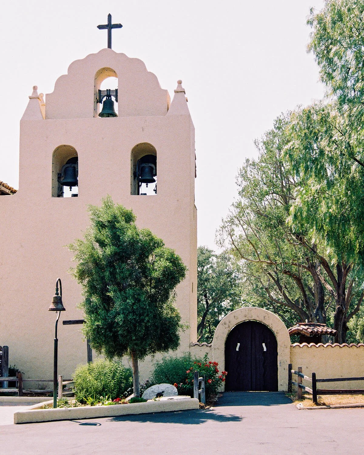 A pink stucco church with a bell tower featuring three bells, a cross on top, a wooden arched door, a small garden with a tree and flowers in front, and a street lamp on the left.
