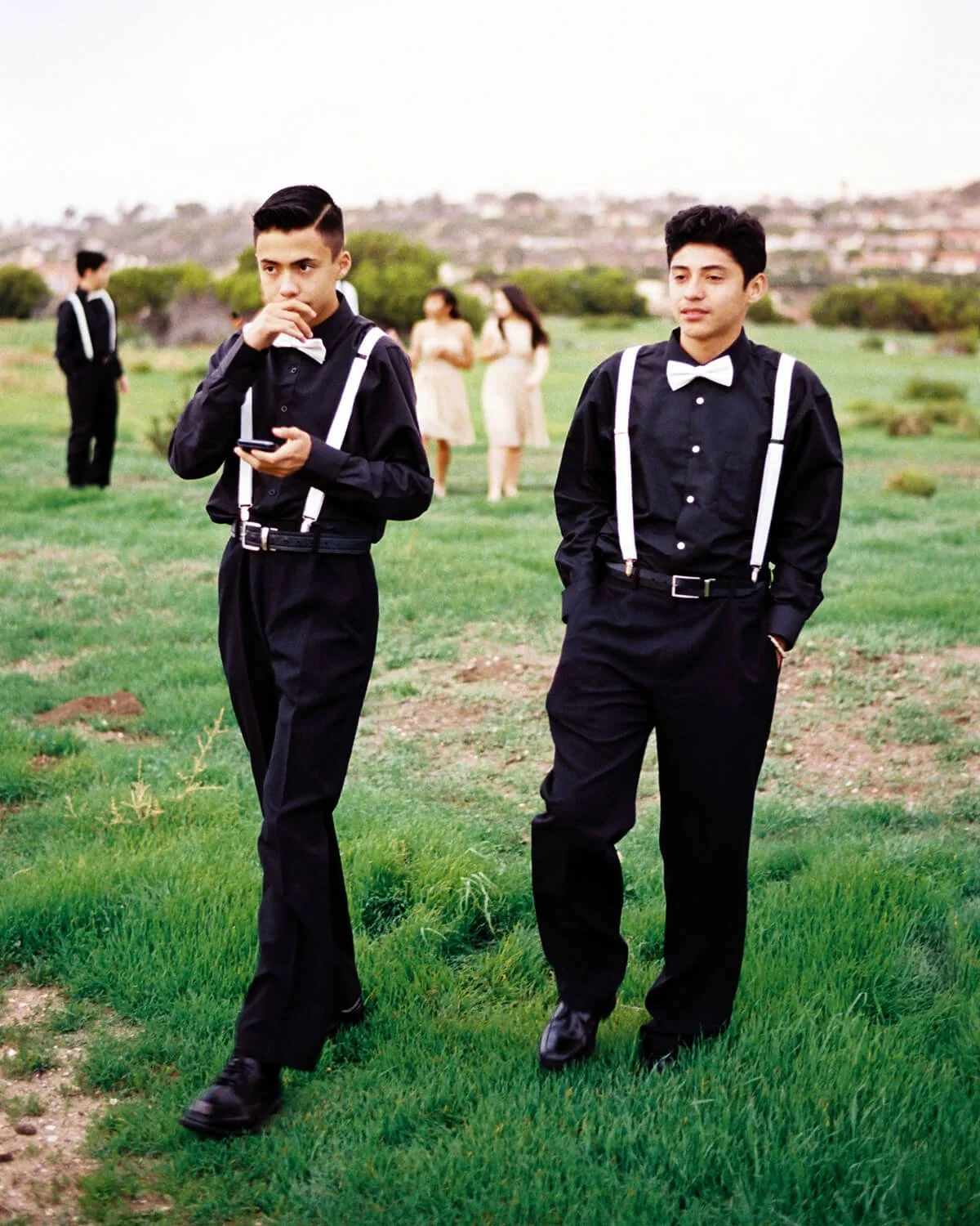 Two chambelanes in black dress shirts, pants, white suspenders, and bow ties walking on grass with two damas in the background.