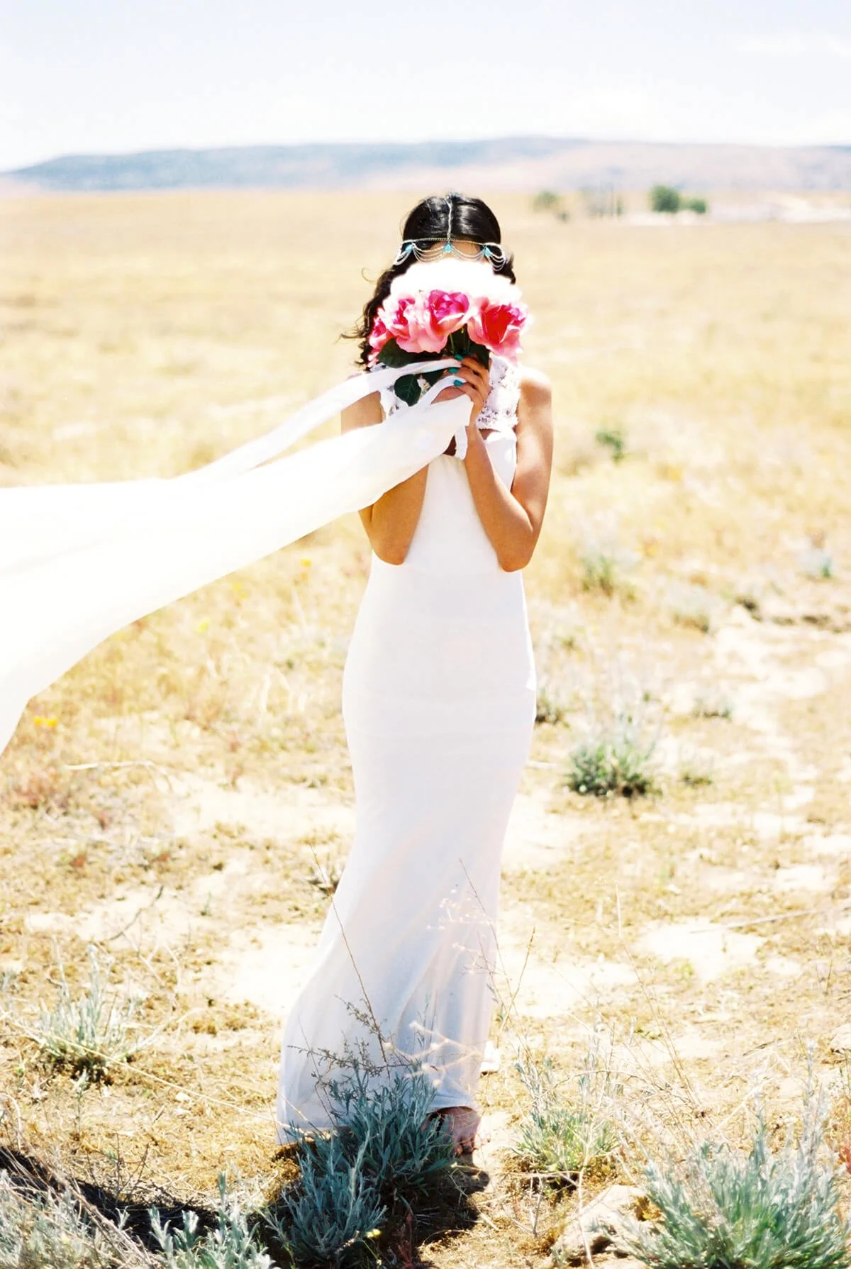 A Quinceañera in a white dress holding a bouquet of pink flowers in front of her face, standing in a dry, open field.