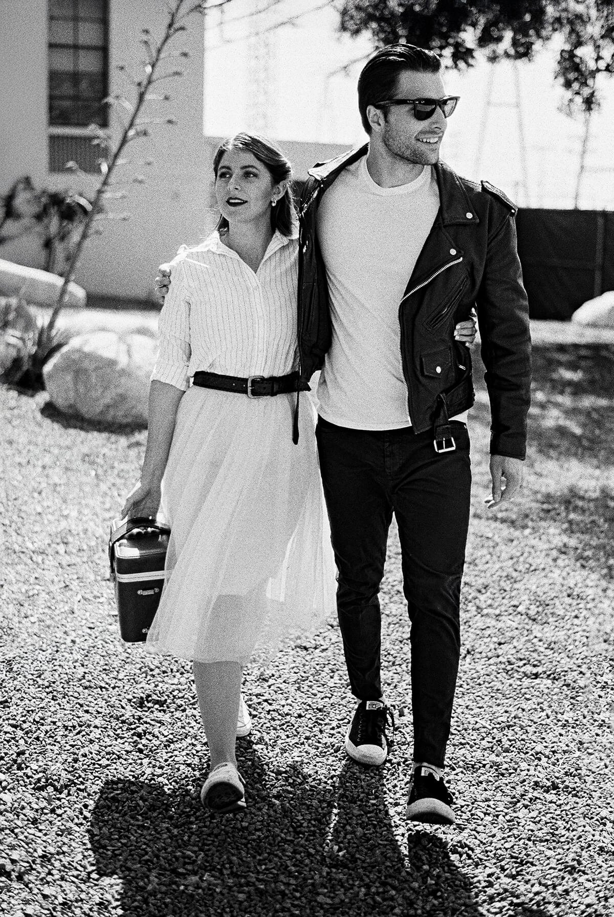 Black-and-white photo of a stylish couple walking outdoors, with the man wearing sunglasses and a leather jacket and the woman carrying a handbag.
