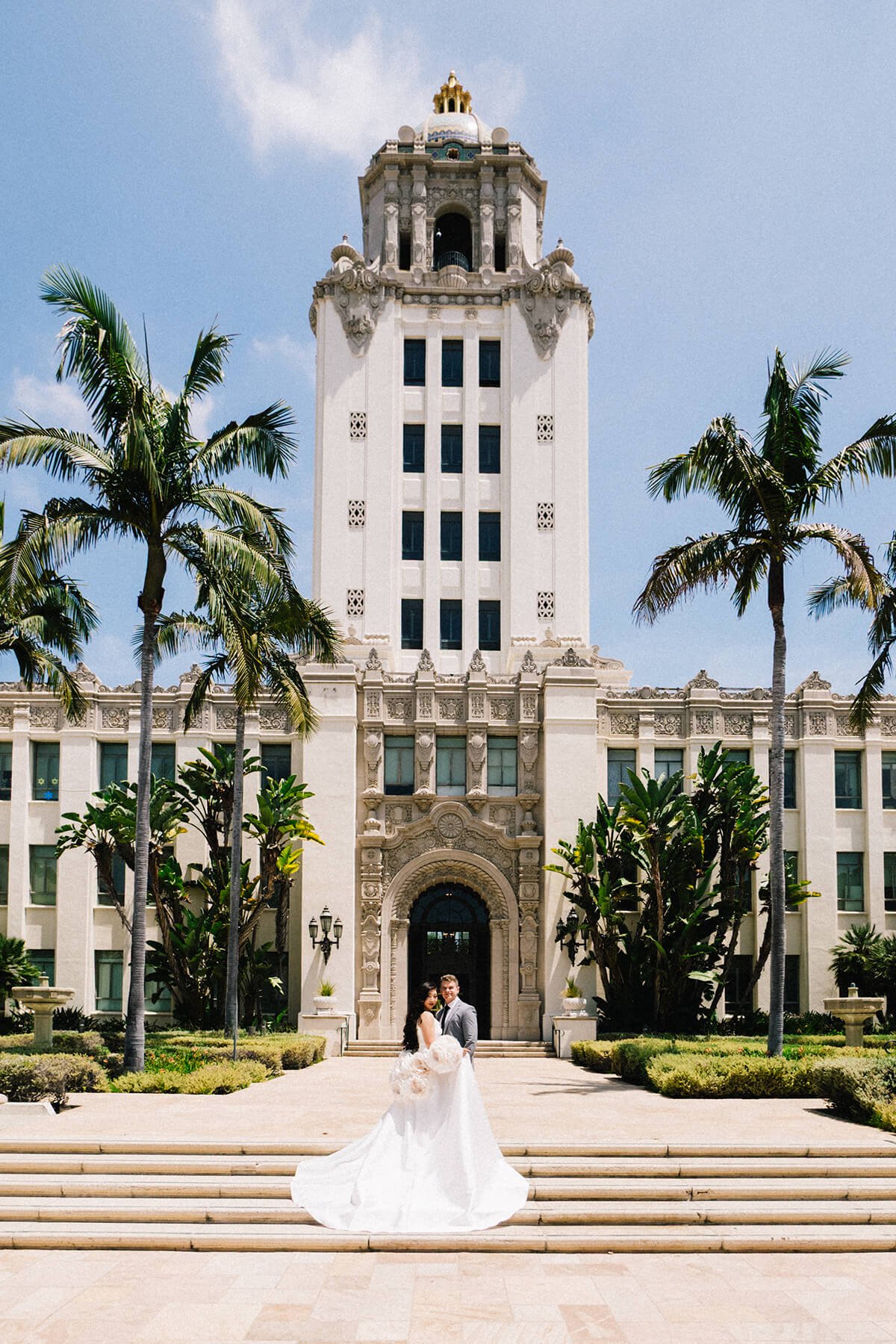 A wedding couple standing in front of the historic Beverly Hills City Hall building with a tall tower, palm trees, and gardens under a clear blue sky.
