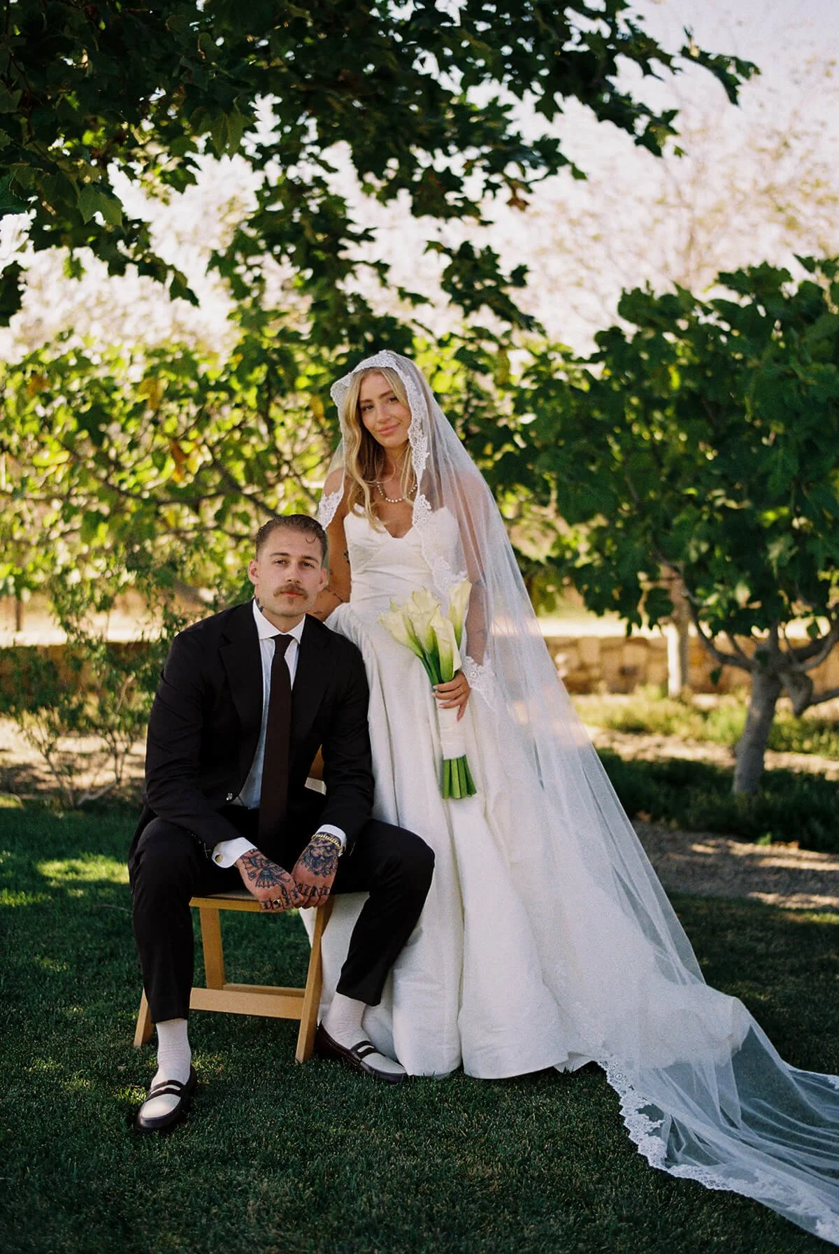 A bride holding a bouquet of calla lilies and a groom dressed in a black suit with tattoos on his hands, both outdoors under lush green trees with flowering branches in the background.