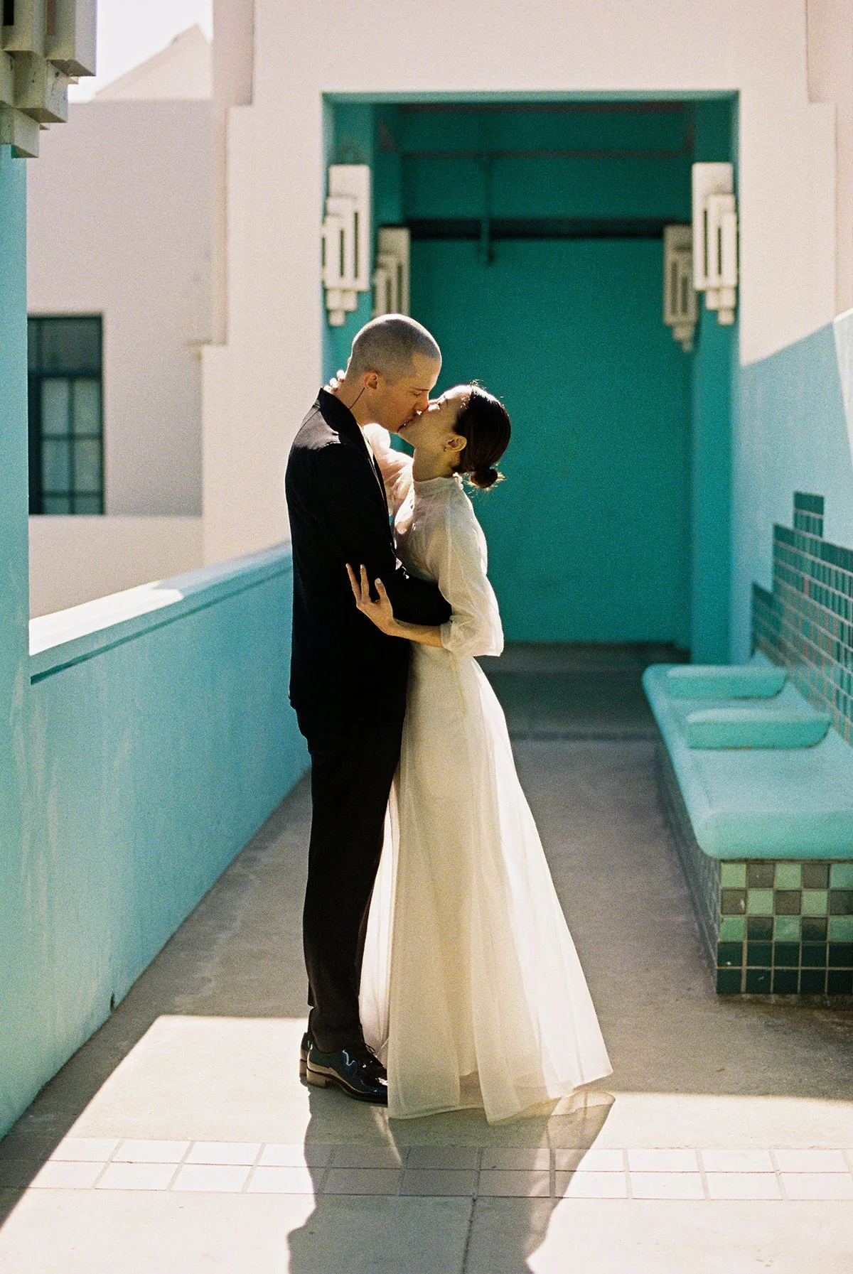 A couple dressed in wedding attire sharing a kiss on a rooftop with teal walls and seating area, during daylight.