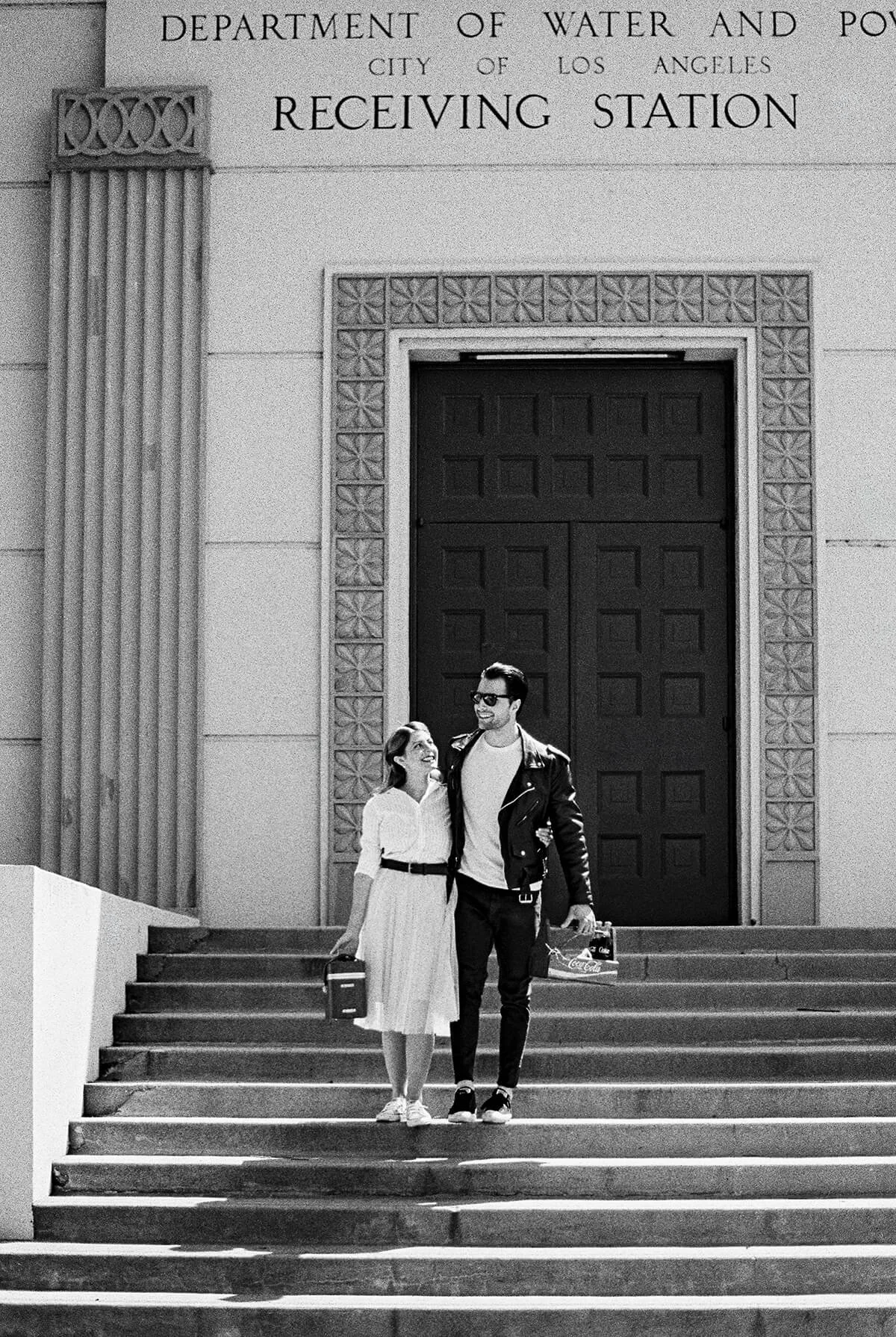 A smiling young woman and man walk hand-in-hand down a staircase in front of a large building with a sign that reads 'Department of Water and Power, City of Los Angeles, Receiving Station.' They are dressed casually, carrying shopping bags and suitca