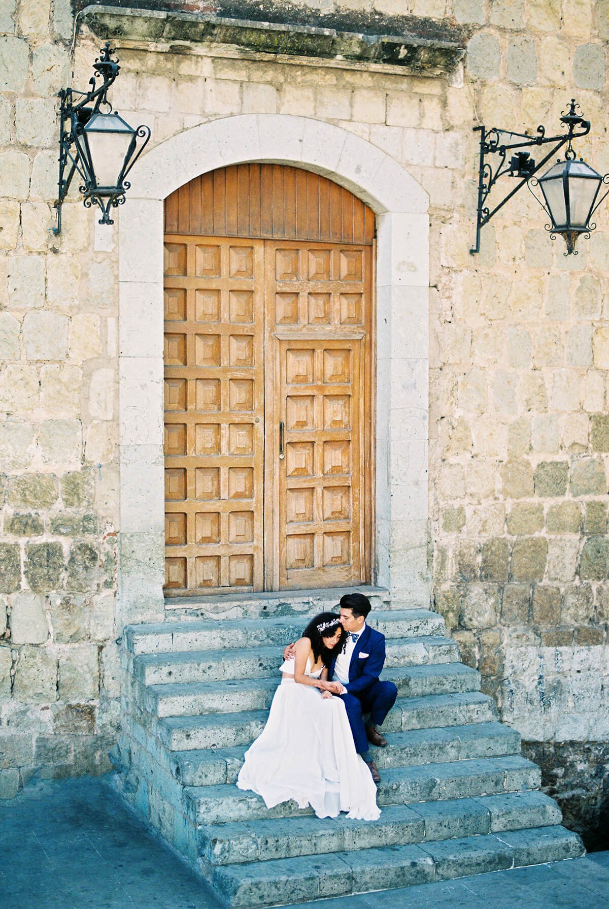 A bride and groom sitting on stone steps in front of a large wooden door, with two black wrought iron lanterns on the stone wall behind them.
